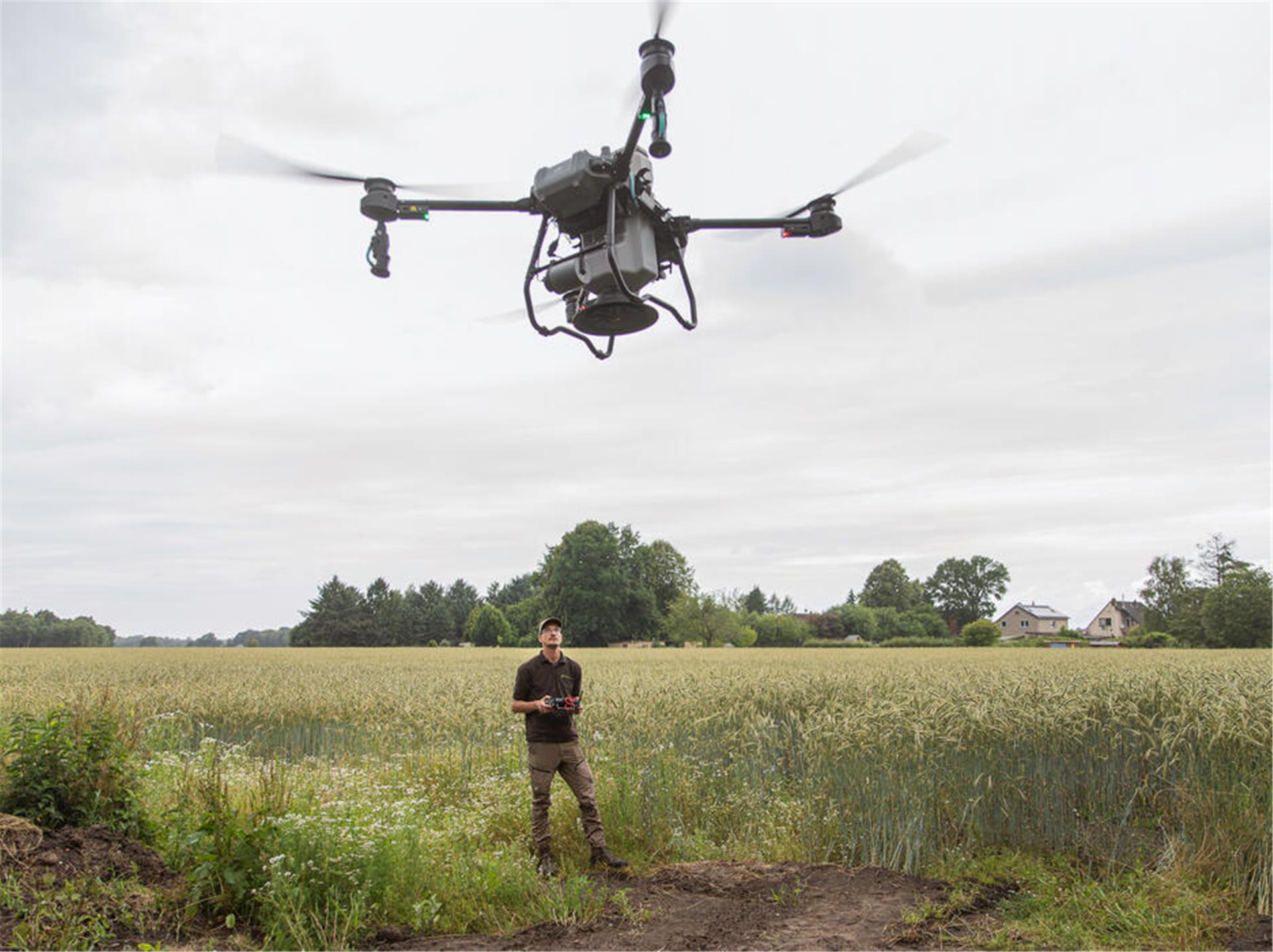 Eine Drohne fliegt durch die Luft. Im Hintergrund steht ein Mann auf dem Feld, der die Drohne steuert.