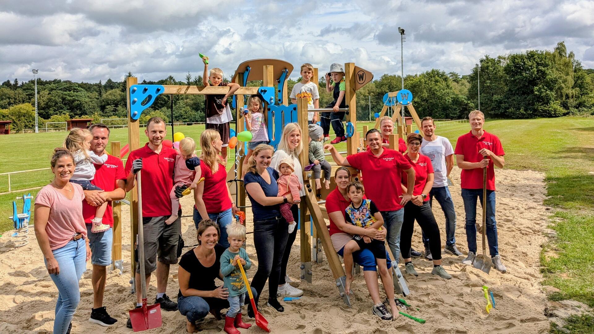 Gruppe von Erwachsenen und Kindern vor einem neu aufgebauten Spielplatz mit Klettergerüst und Rutsche auf einem Sandplatz, im Hintergrund ein Sportplatz und Bäume. Einige tragen rote Vereinsshirts, mehrere Kinder spielen oder werden getragen.