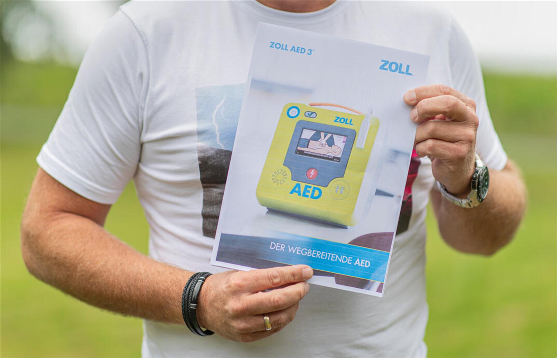Ein Zettel, auf dem ein Defibrillator abgebildet ist, wird von einem Mann in die Kamera gehalten. Zu sehen sind nur die Arme und der Bau des Mannes, alles andere ist "abgeschnitten".