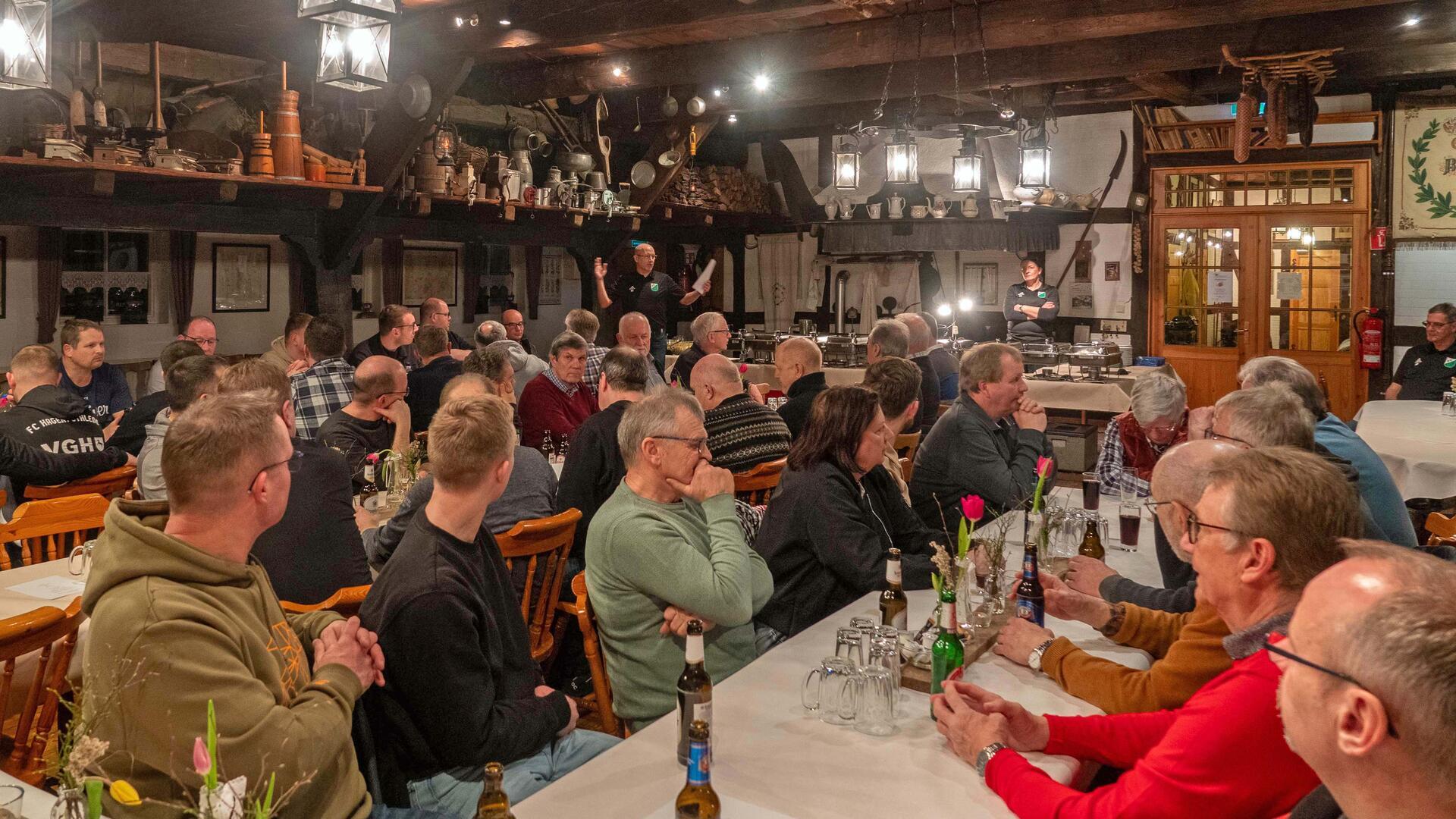 Ein geselliges Treffen in einer rustikalen Gaststube mit vielen Personen, die an langen Tischen sitzen. Eine Person steht vorne und hält eine Rede, während die Gäste aufmerksam zuhören. Die Wände und Decken sind mit alten Haushaltsgegenständen dekoriert, und die Beleuchtung besteht aus traditionellen Laternen. Auf den Tischen stehen Getränke und Blumendekorationen.