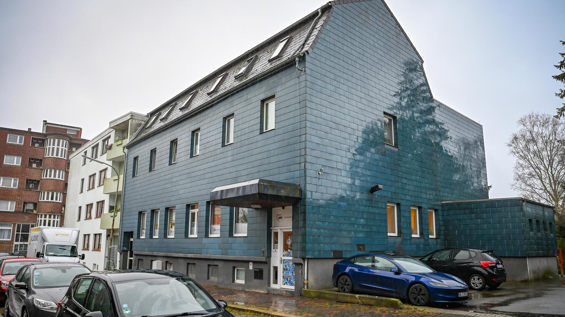 Mehrfamilienhaus mit blau-grauer Fassade im Regen, davor und daneben geparkte Autos.