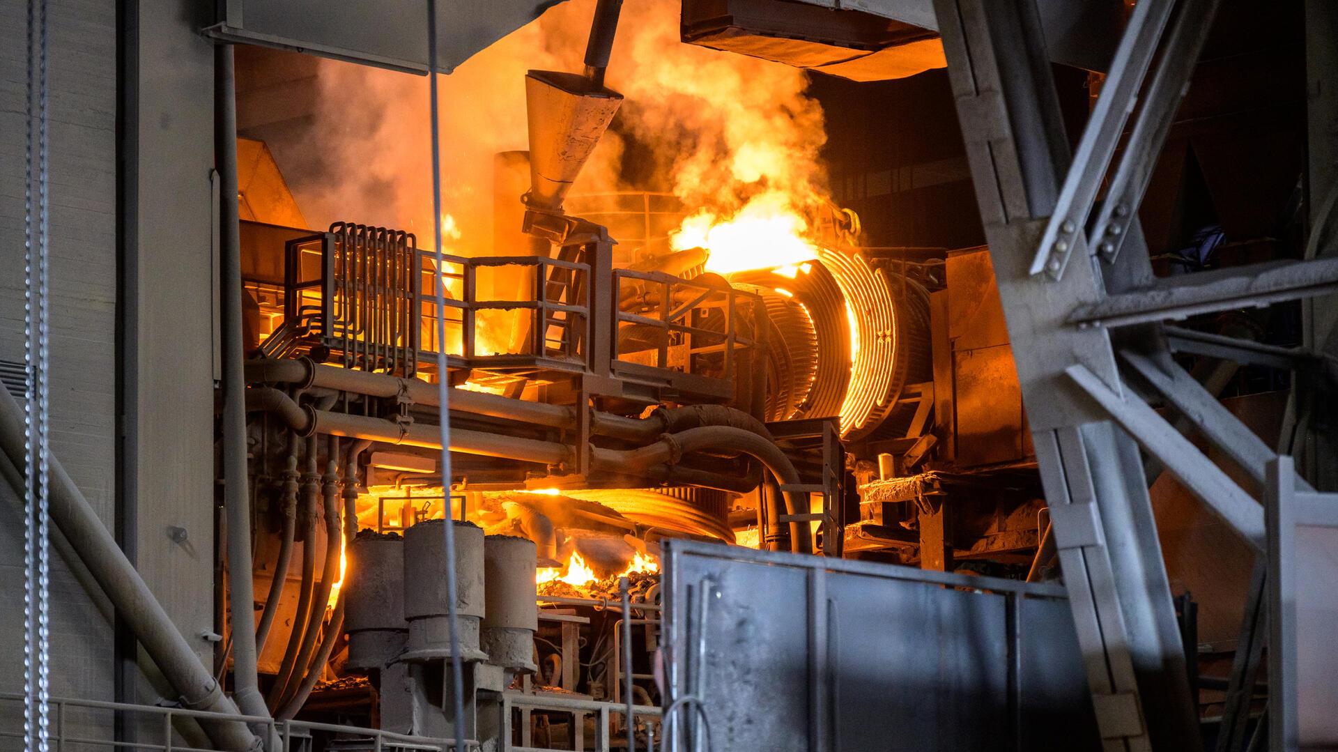 Blick ins Innere eines Stahlwerks: Glühende, orangefarbene Flammen und flüssiges Metall schieben sich durch Rohre und Walzen, umgeben von massiven Stahlträgern und Industrieanlagen.