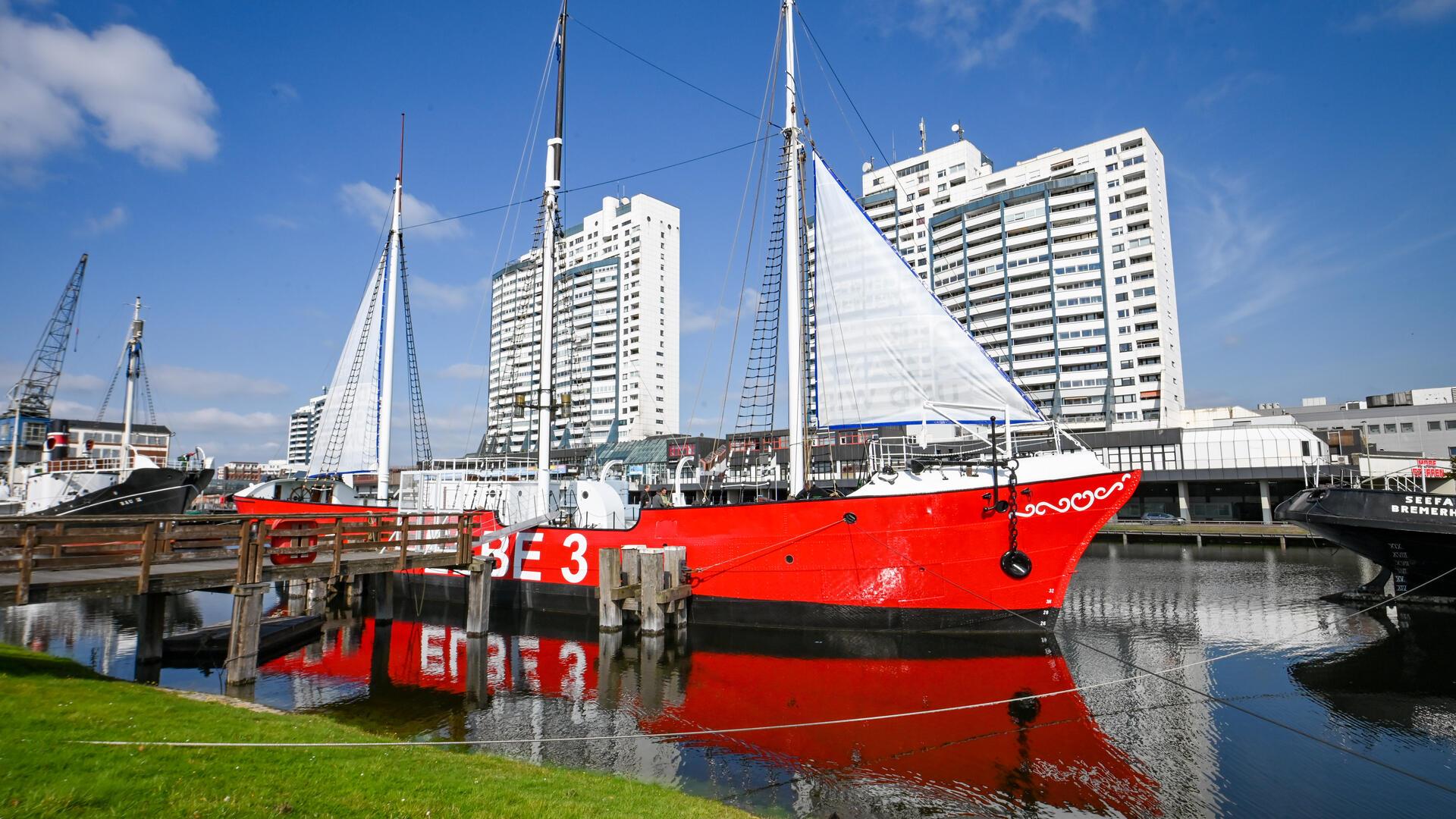 Historisches Feuerschiff ELBE 3 mit roten Rumpf und weißen Segeln im Alten Hafen Bremerhaven, umgeben von weiteren Museumsschiffen und Hochhäusern im Hintergrund.