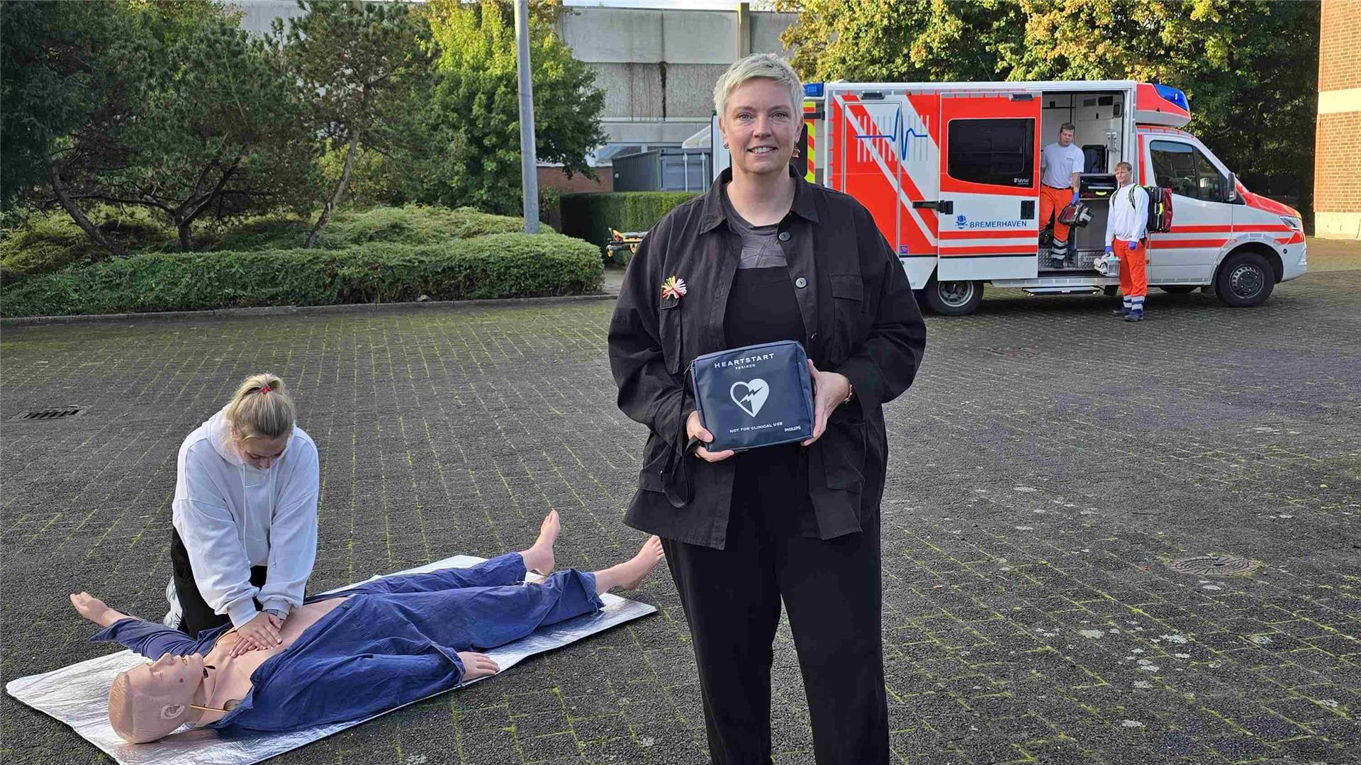 Eine Frau hält einen Defibrillator in den Händen, hinter ihr übt eine junge Frau an einer Reanimationspuppe die Herz-Druckmassage. Im Hintergrund steht ein Rettungswagen der Feuerwehr Bremerhaven mit zwei Einsatzkräften.