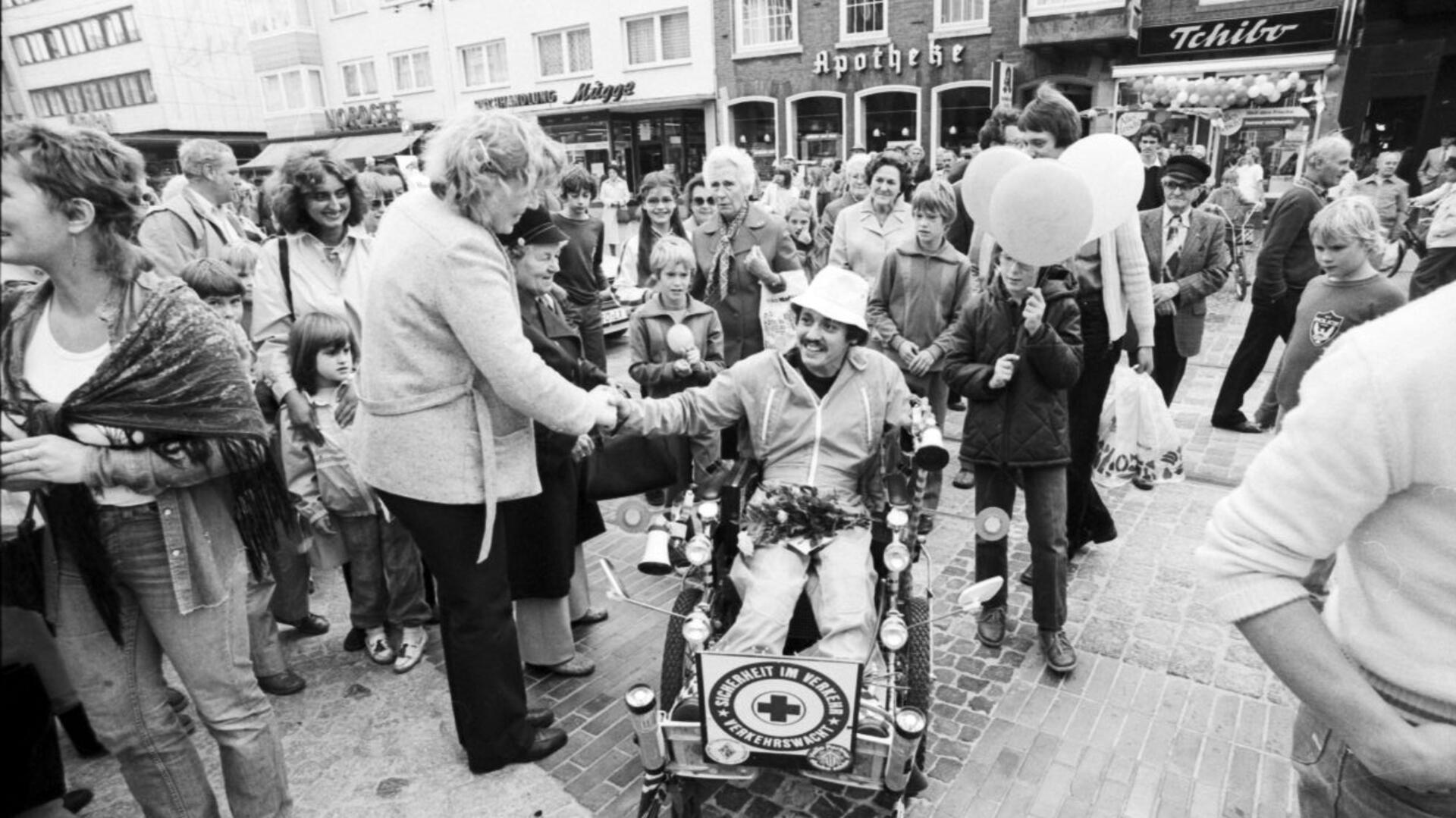 Schwarz-Weiß-Foto zeigt Rollstuhlfahrer umringt von Menschen. Einige gratulieren.