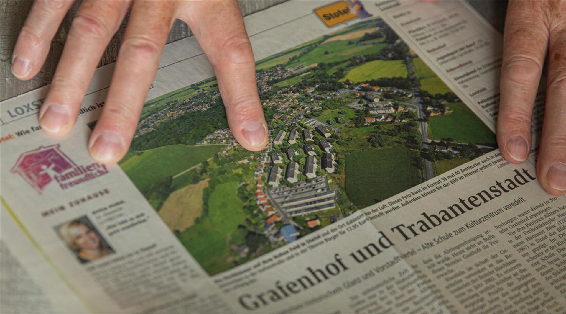 Ein Foto von einem Wohnviertel, das in der Zeitung erschien. Zwei Hände liegen auf der Zeitung, ein Finger deutet auf das Foto.