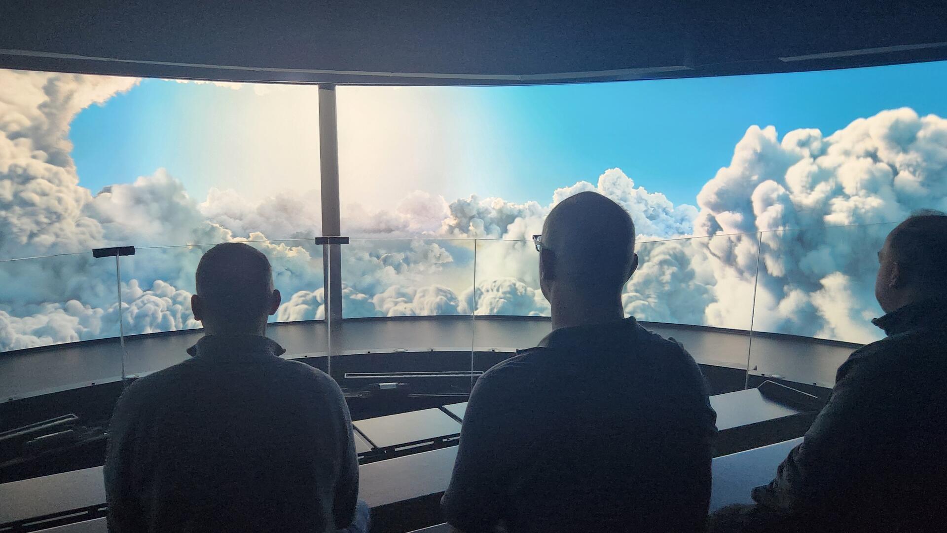 Drei Menschen stehen vor einer Leinwand und betrachten Wolken und Himmel. Die neue Dauerausstellung "Wetterextreme" wird jetzt für die Öffentlichkeit eröffnet. 
