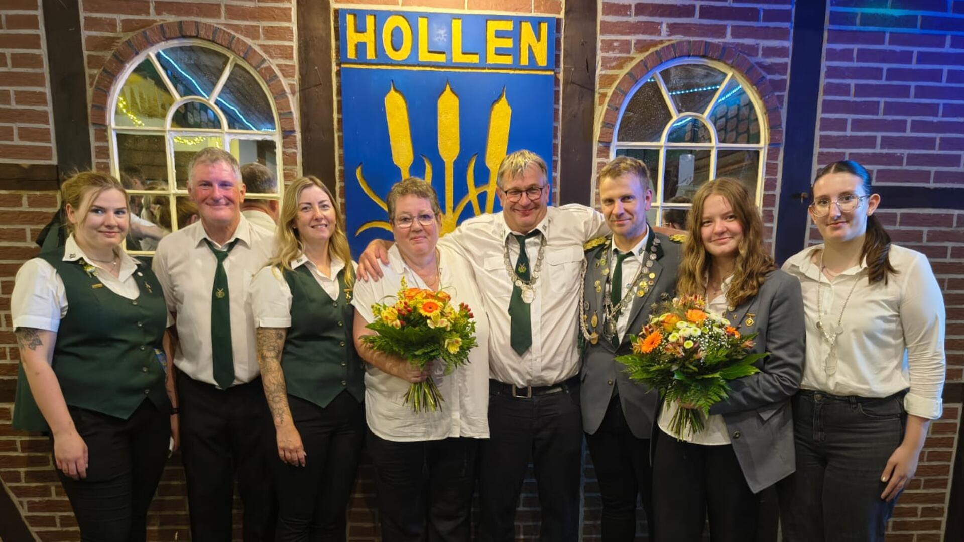 **ALT-Text:**
Gruppenfoto des neuen Königshauses des Schützenvereins Hollen: Acht Personen, teils in Uniform, stehen vor einem Schild mit der Aufschrift „Hollen“, zwei von ihnen halten Blumensträuße.