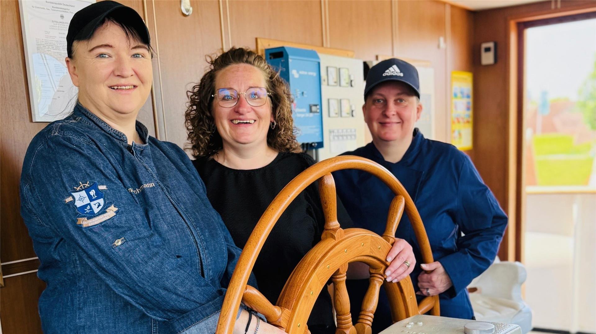 Drei Frauen stehen lächelnd an einem hölzernen Steuerrad auf der "MS Hansa"