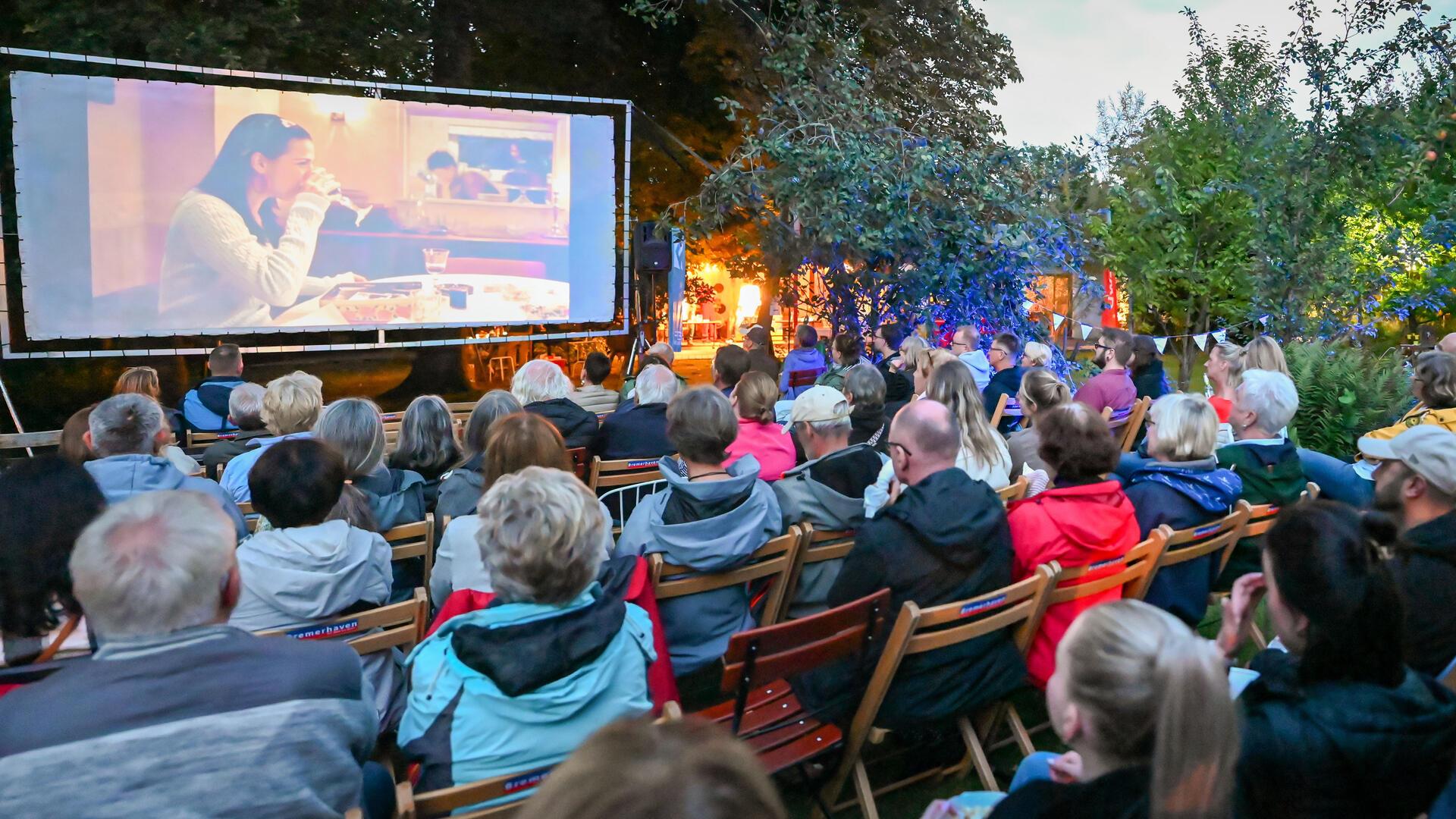 Menschen sitzen auf Stühlen im Freien vor einer Leinwand.