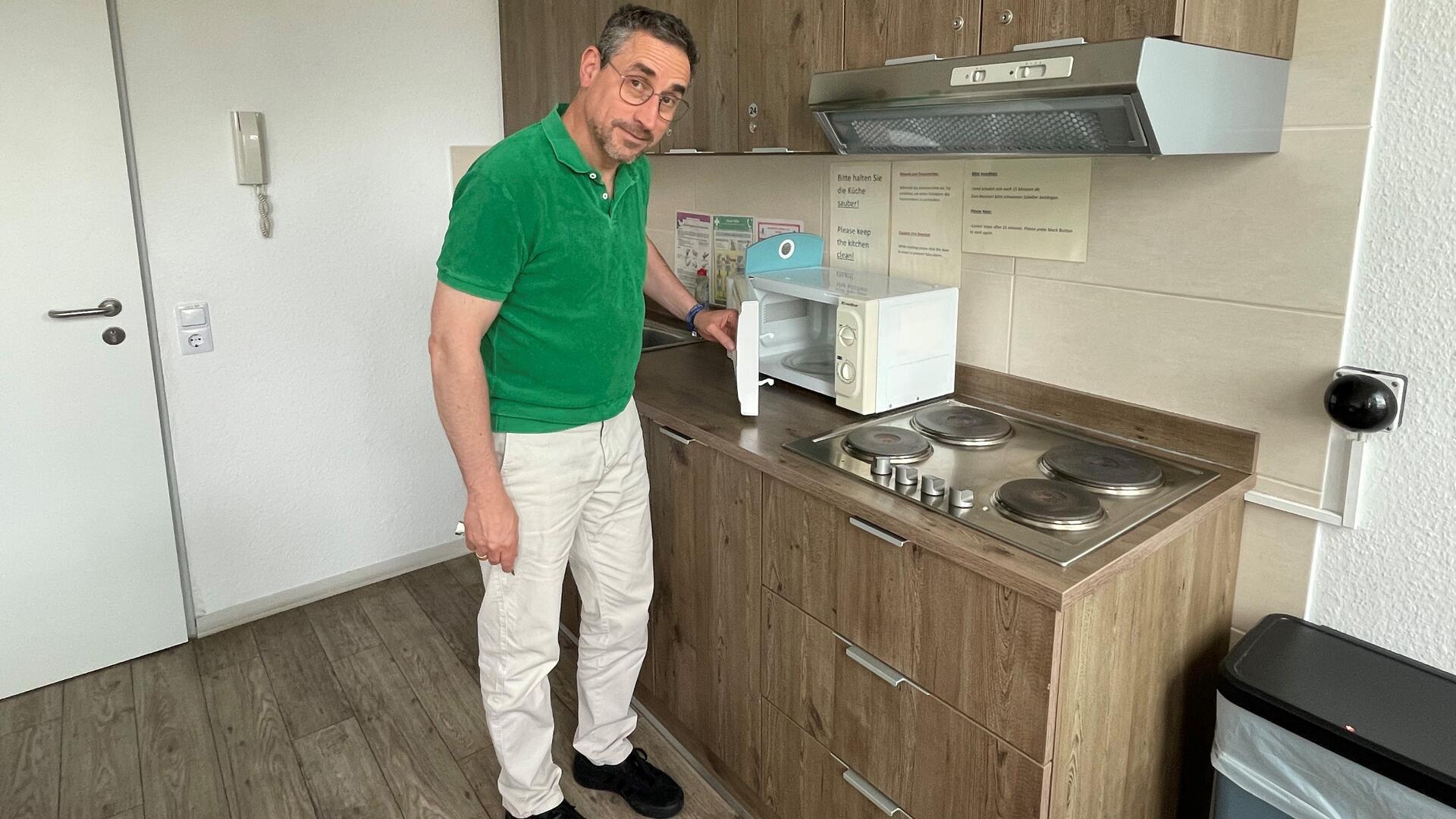 Ein Mann mit Brille, grünem Polo-Hemd und weißer Hose steht in einer Küche. Er öffnet die Mikrowelle, die links neben dem Herd steht. Darüber die Abzugshaube. Die Küche besteht aus hellbraunen Holzschränken.