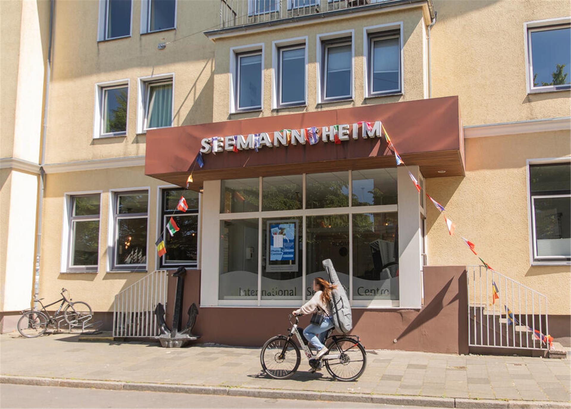 Ein gelbes Gebäude, an dem eine Frau auf einem Fahrrad vorbeifährt. Über dem Eingang steht "Seemannsheim".