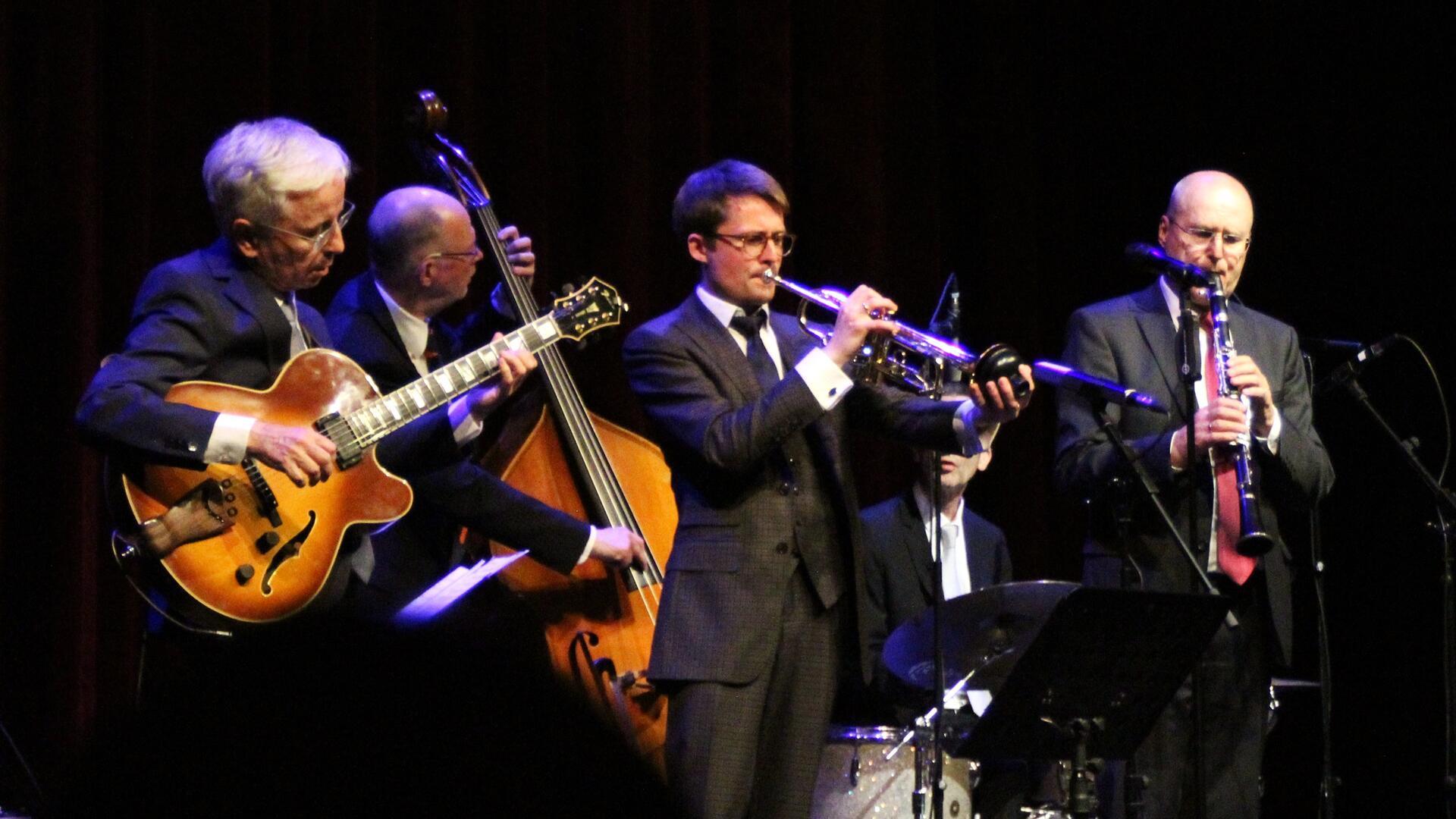 Vier Jazzmusiker auf einer Bühne: Ein Gitarrist, ein Kontrabassist, ein Trompeter und ein Klarinettist spielen gemeinsam in eleganter Abendkleidung, umgeben von Mikrofonen und Instrumenten.
