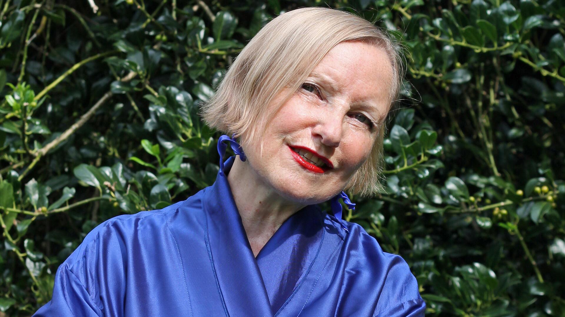 Eine Frau in einem blauen Outfit vor grünem Laub, lächelnd mit rotem Lippenstift. Es ist Anna Haentjens, die im TiF Bremerhaven auftreten wird - mit einer Hommage an Lale Andersen. 