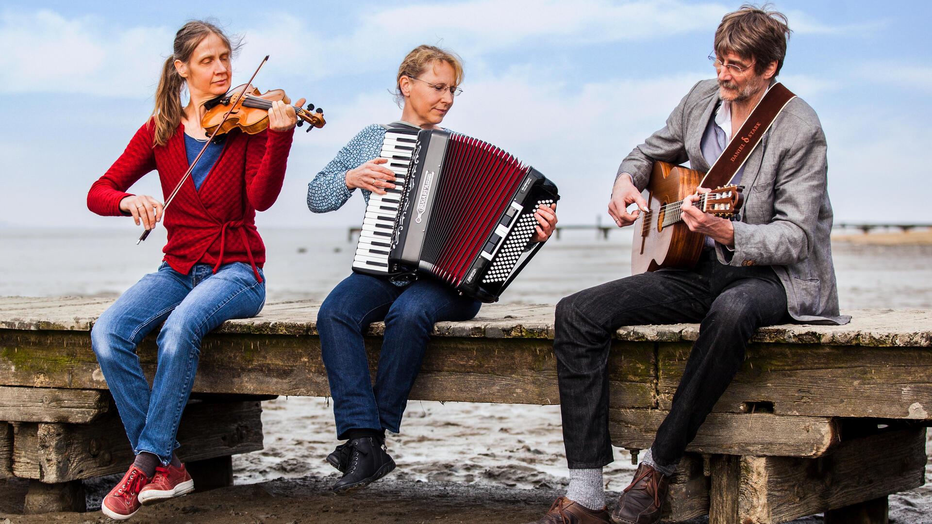 Drei Musiker sitzen mit ihren Musikinstrumenten auf eiem Steg. 