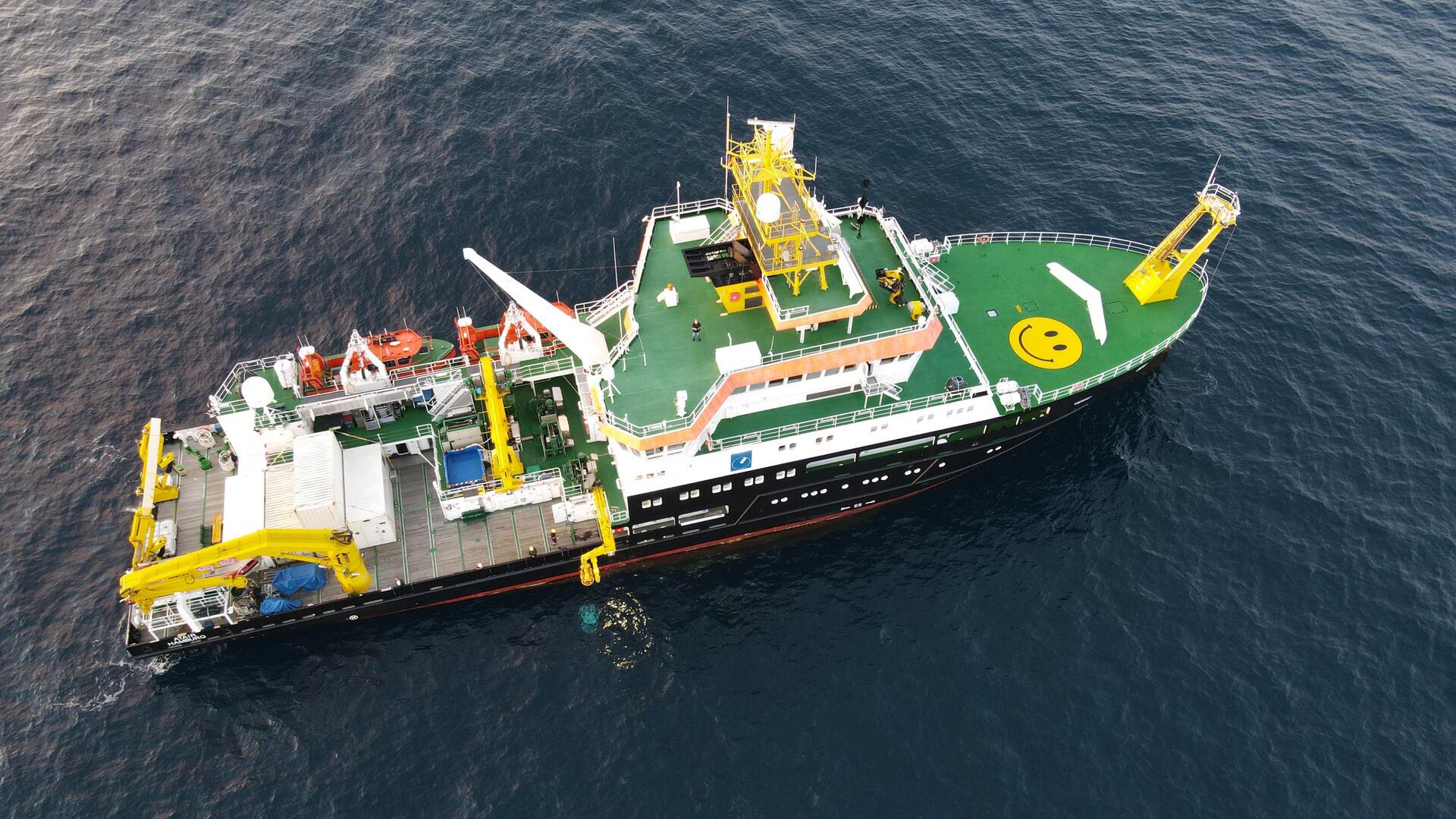 Forschungsschiff ATAIR des Bundesamts für Seeschifffahrt und Hydrographie (BSH) von oben auf offener See, mit grünem Helikopterdeck, gelbem Smiley und wissenschaftlicher Ausrüstung an Bord.