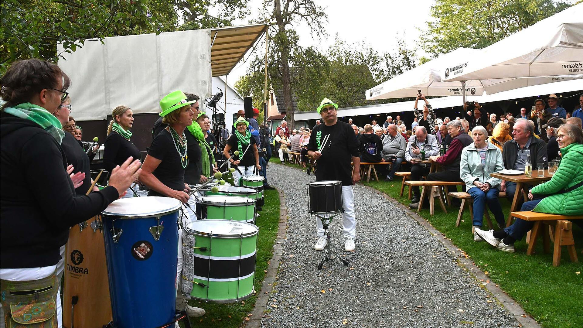 Eine Trommelgruppe mit grünen Accessoires tritt beim letzten Grilleabend in Wremen auf. Vor der Bühne sitzen zahlreiche Gäste an Bierbänken im Kurgarten.