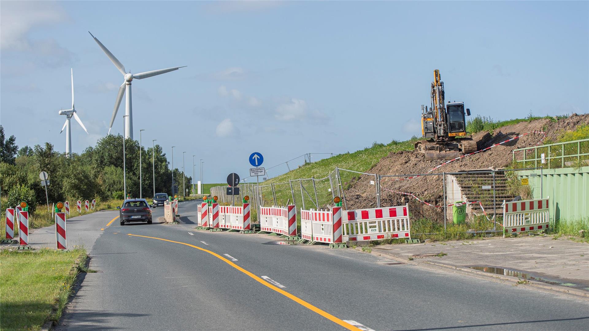 Eine Straße, ein Bagger auf einem Deich und zwei Windräder im Hintergrund.