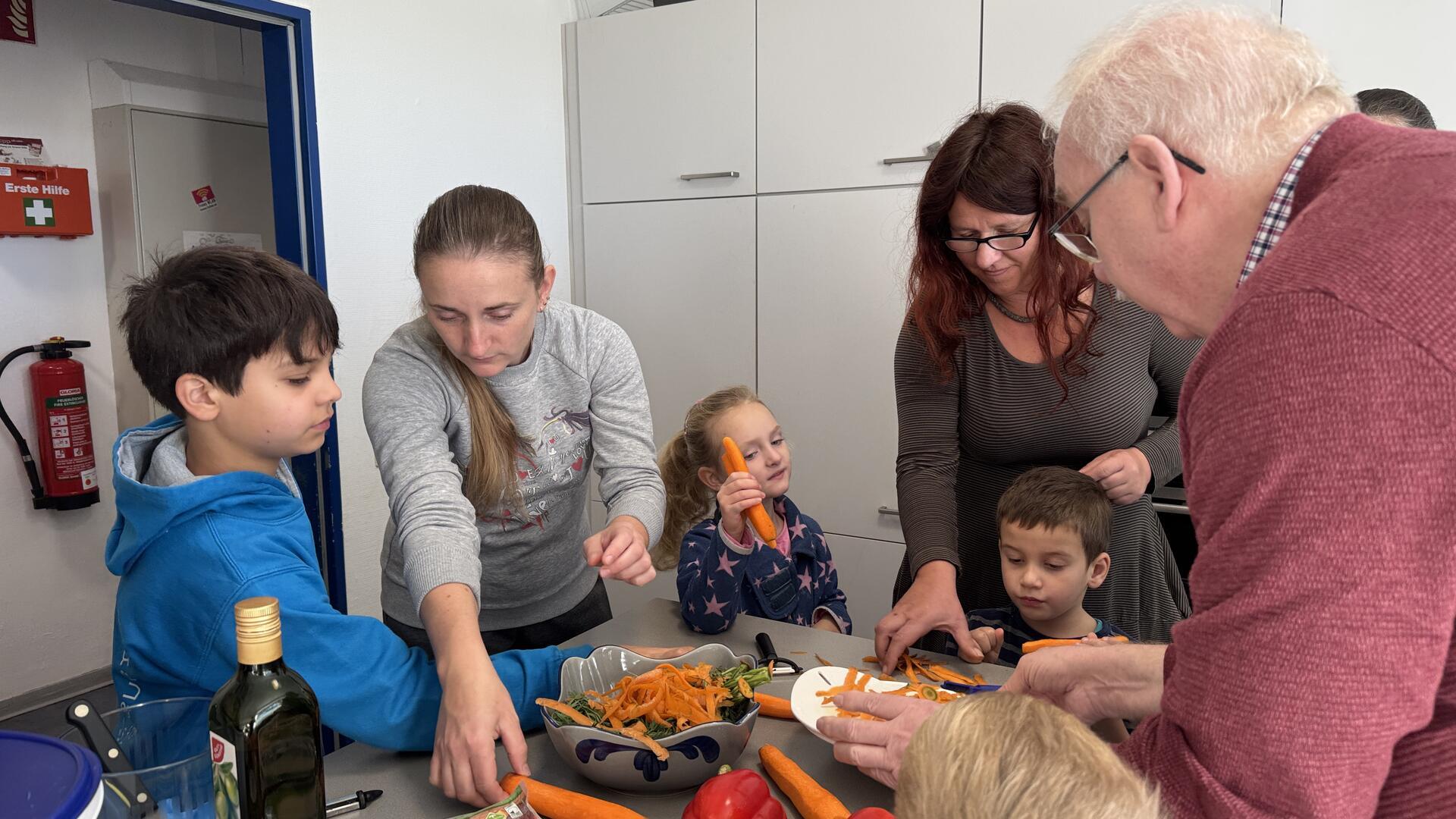 „Allein*erziehend vernetzt“ bringt Generationen an einen Tisch Mehrere Kinder, Erwachsene und ein älterer Mann schälen gemeinsam Karotten an einem Tisch in einer Küche. Sie wirken konzentriert und fröhlich beim gemeinsamen Kochen.