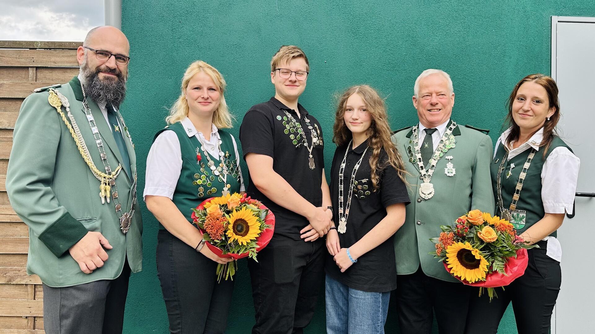 Gruppenfoto der neuen Schützenhoheiten des Schützenvereins Neuenlande. Drei Männer und drei Frauen tragen Schützenketten, zwei Frauen halten bunte Blumensträuße mit Sonnenblumen. Alle stehen vor einer grünen Wand und lächeln in die Kamera.