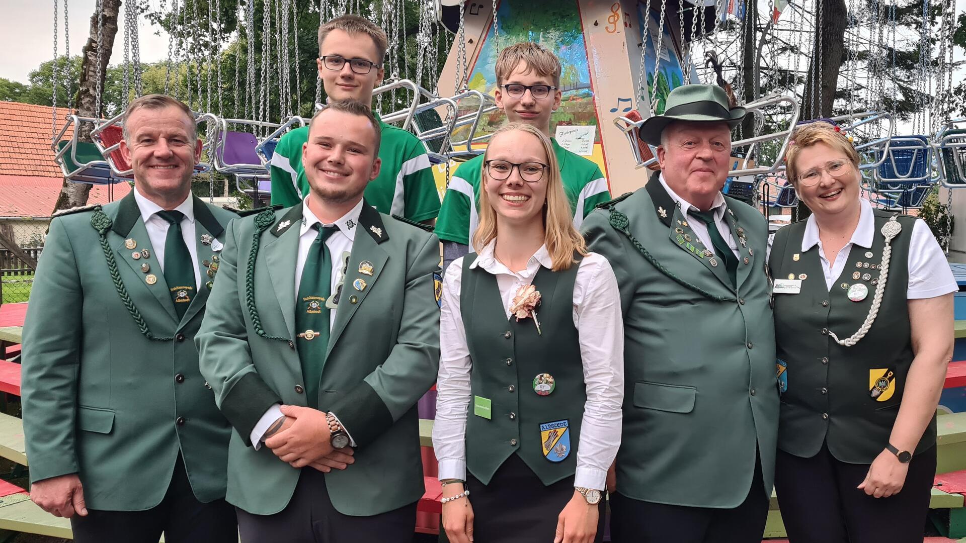 Eine Gruppe von acht Personen, bestehend aus Männern und Frauen, posiert lächelnd für ein Gruppenfoto vor einem Karussell. Die vordere Reihe trägt traditionelle grüne Uniformen und Westen mit Abzeichen und Schützenorden, während zwei junge Männer in der hinteren Reihe grün-weiße Sportshirts tragen.