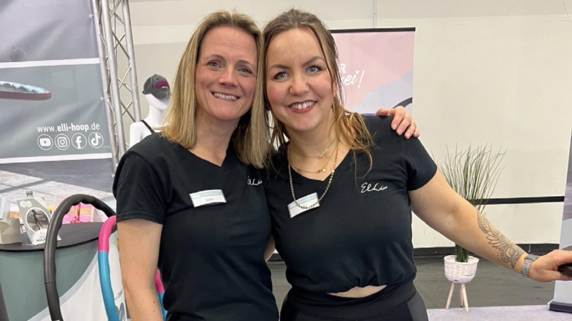 Zwei Frauen stehen lächelnd nebeneinander an einem Messestand, tragen schwarze T-Shirts mit Namensschildern und dem Aufdruck „Elli“. Im Hintergrund sind Hula-Hoop-Reifen und Werbematerialien zu sehen.