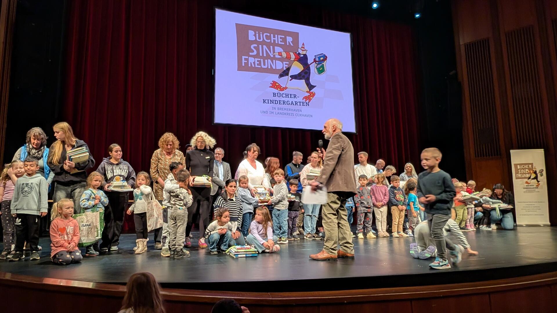 Kinder und Erwachsene stehen auf einer Theaterbühne. Im Hintergrund ist eine Leinwand mit dem Logo „Bücher sind Freunde“ zu sehen.