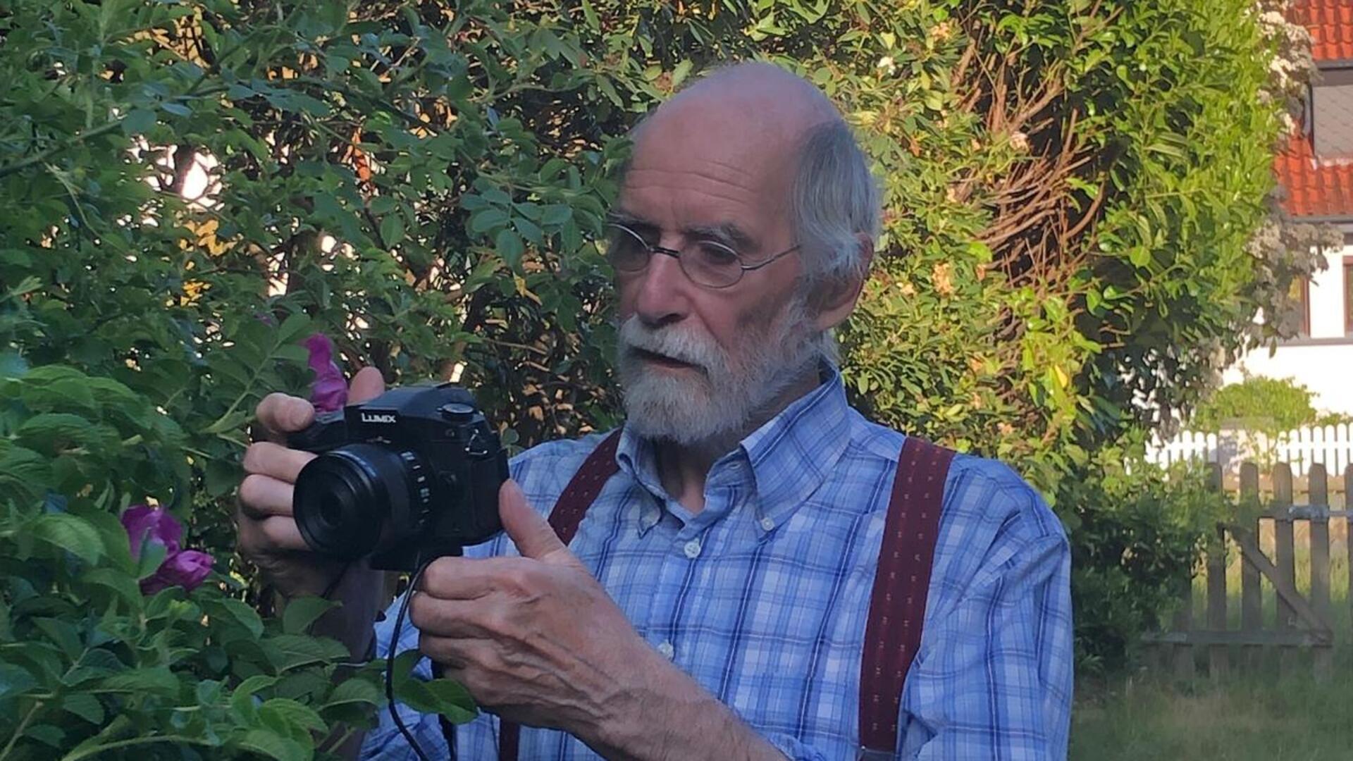 Ein älterer Mann mit Brille und grauem Bart fotografiert mit einer Kamera Pflanzen im Garten.