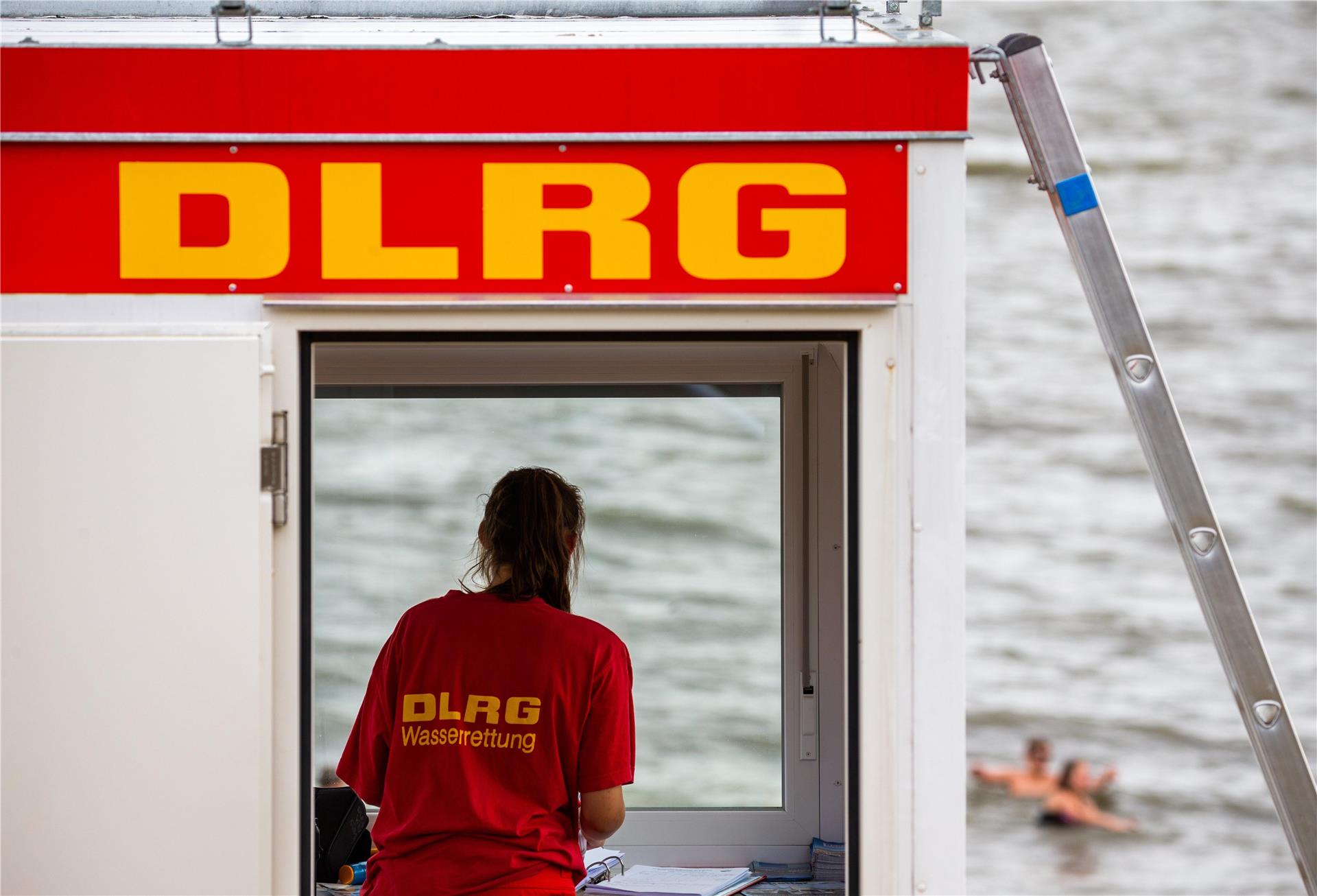 Ein Teil eines DLRG-Häuschens ist zu sehen, sowie eine Frau, die ein rotes Shirt trägt, auf dem in gelber Schrift die Worte "DLRG" und "Wasserrettung" zu lesen sind. Im Hintergrund ist unscharf Wasser zu erkennen sowie Menschen, die dort zu schwimmen scheinen.  