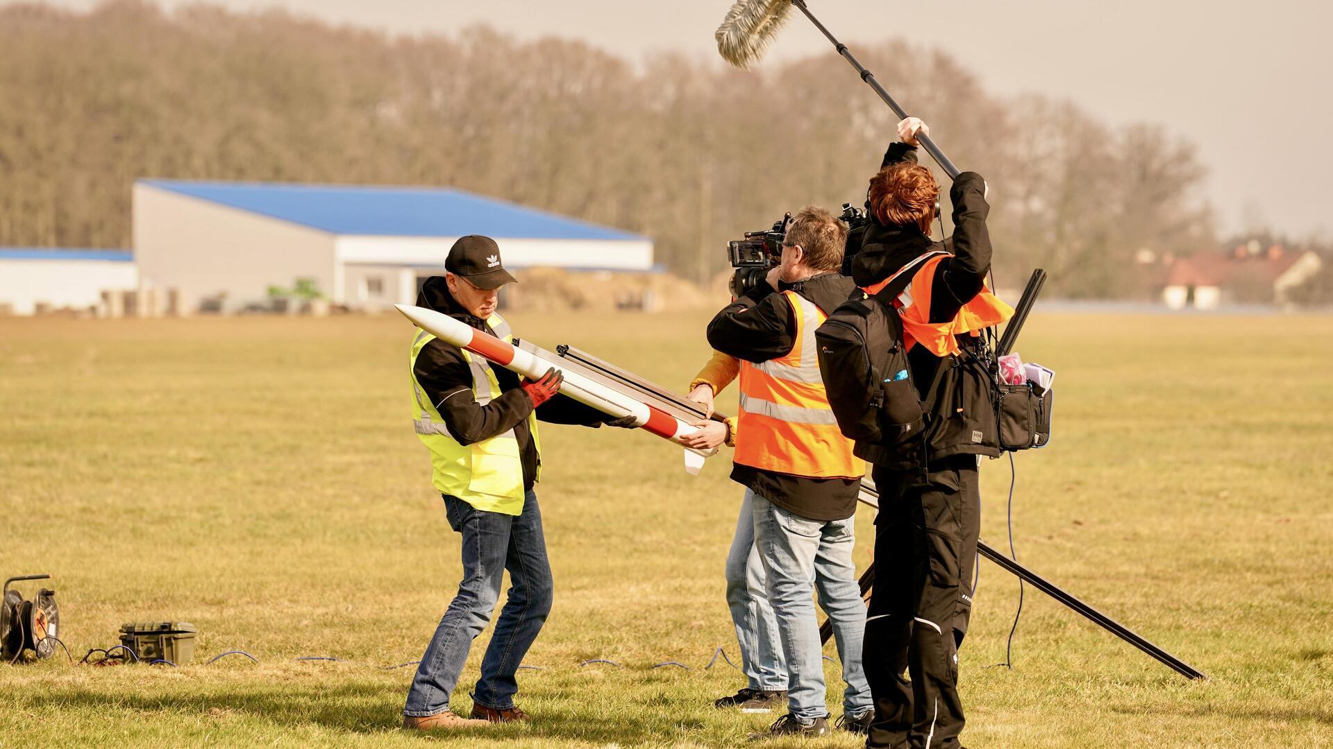 Ein Mann hat eine Rakete in der Hand, dabei wird er von einem Filmteam gefilmt.