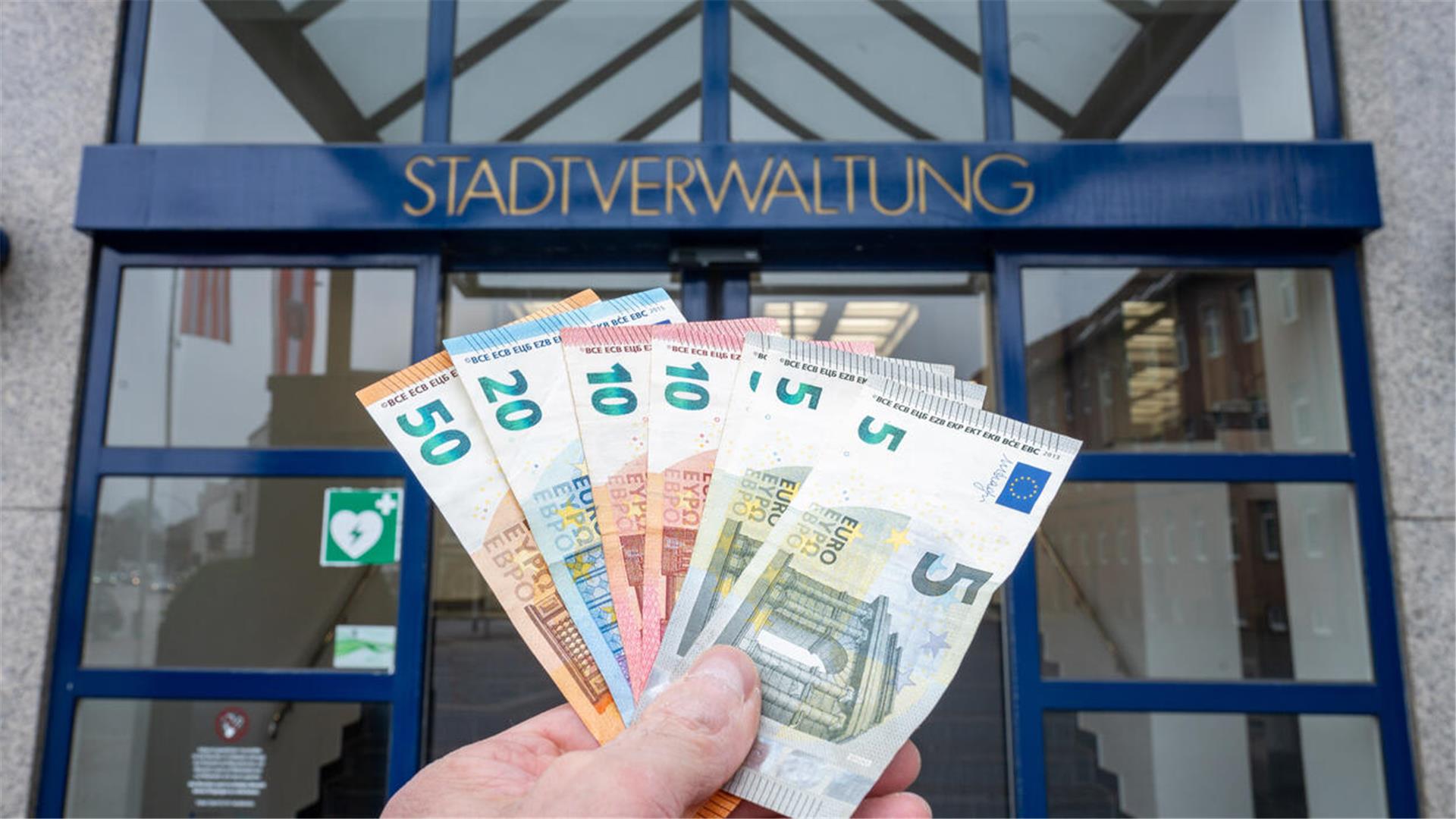 Eine Hand hält Geldscheine vor einer Tür, über der Stadtverwaltung geschrieben steht.