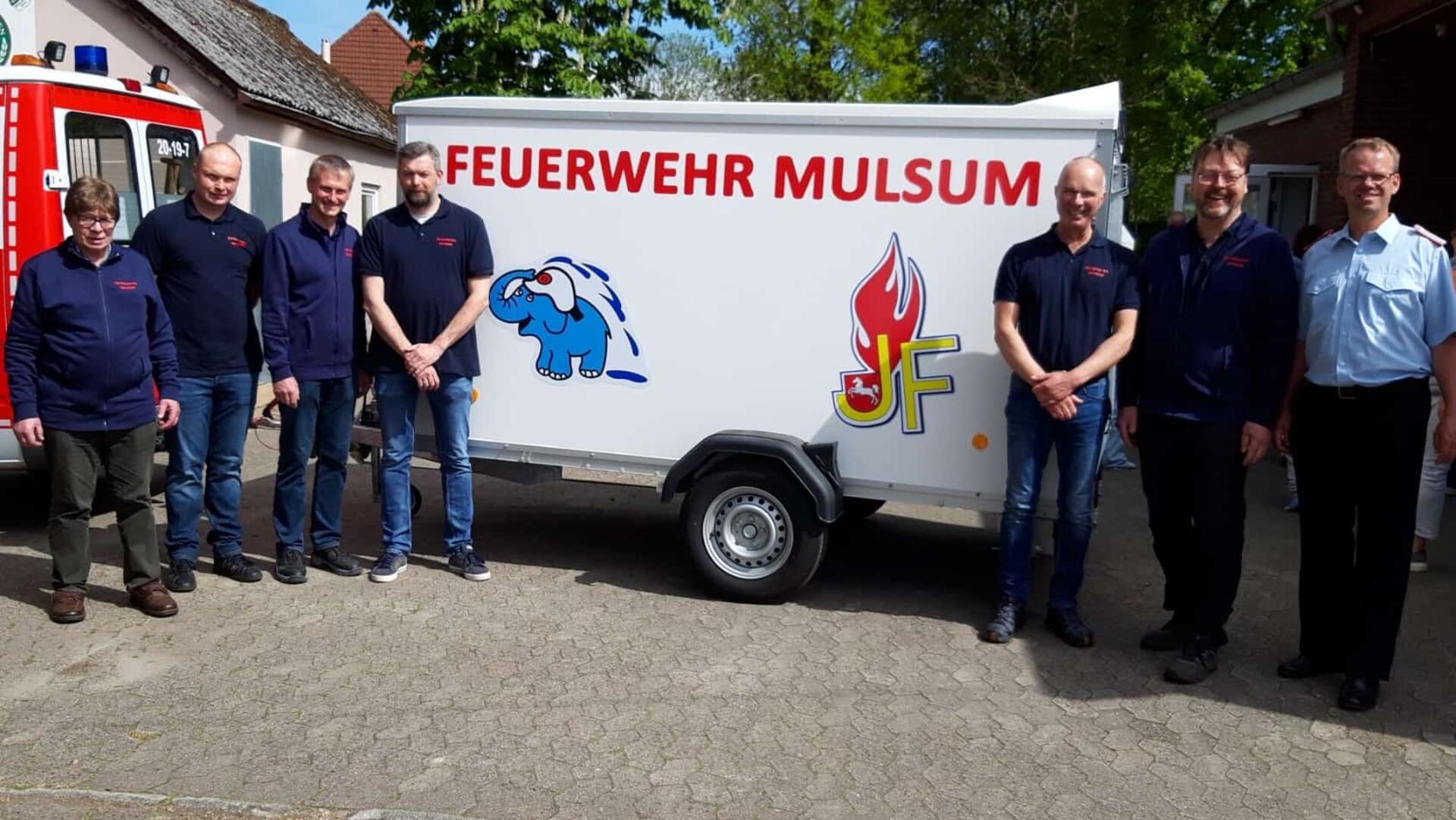 Mehrere Männer stehen vor einem Anhänger mit der Aufschrift „Feuerwehr Mulsum“ und den Logos der Jugend- und Kinderfeuerwehr; links ein Feuerwehrfahrzeug.