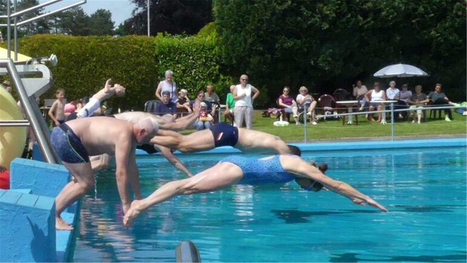 Mehrere Badegäste springen gleichzeitig ins Freibad Midlum, während Zuschauer im Hintergrund auf Stühlen das Geschehen verfolgen.