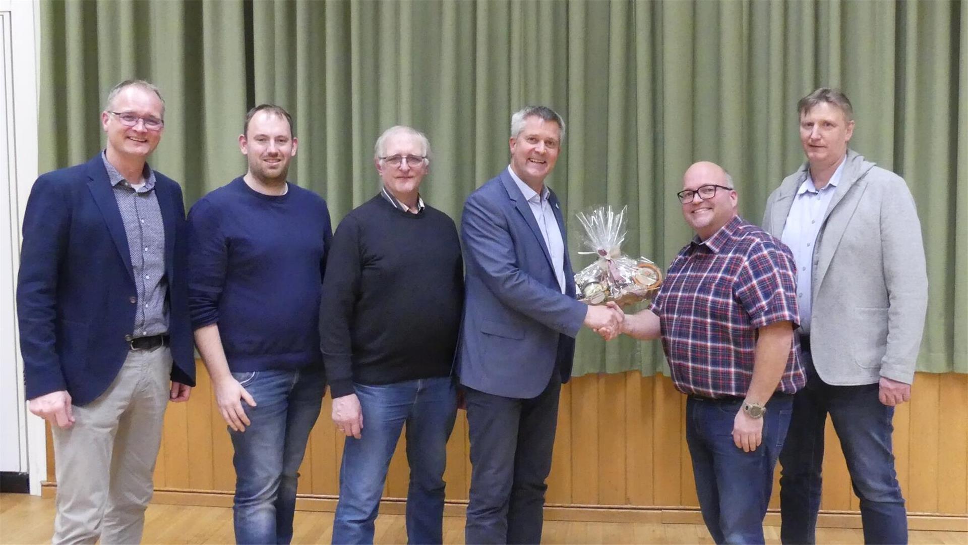 Eine Gruppe von sechs Männern steht nebeneinander in einem Raum mit Holzboden und grünem Vorhang im Hintergrund. Zwei Männer in der Mitte schütteln sich die Hand; einer von ihnen überreicht ein Geschenk.