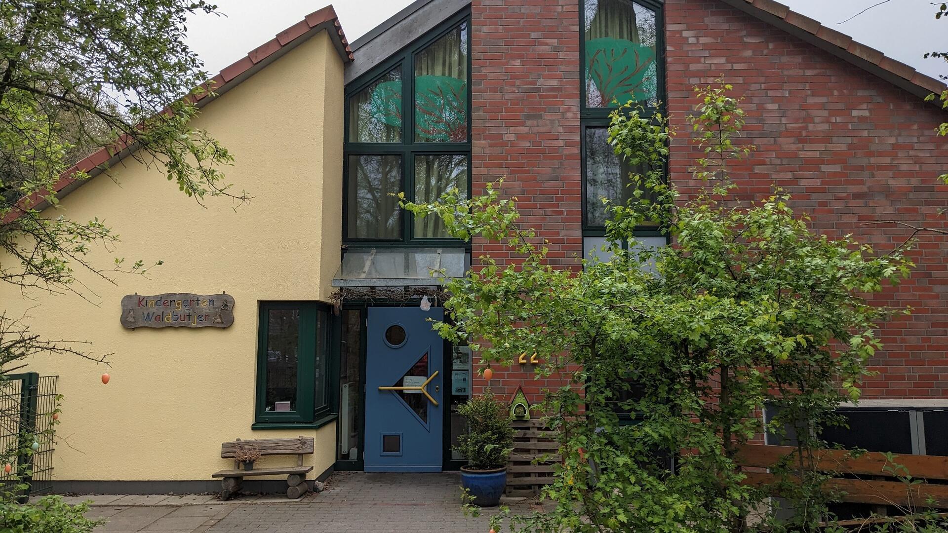 Außenansicht des Kindergartens Waldbutjer. Das Gebäude hat eine gelbe und eine rote Backsteinfassade mit großen Fenstern, die grüne Pflanzenmotive zeigen. Vor dem Eingang steht eine blaue Tür mit einem runden Fenster. Links neben der Tür befindet sich eine kleine Holzbank und ein grüner Strauch.