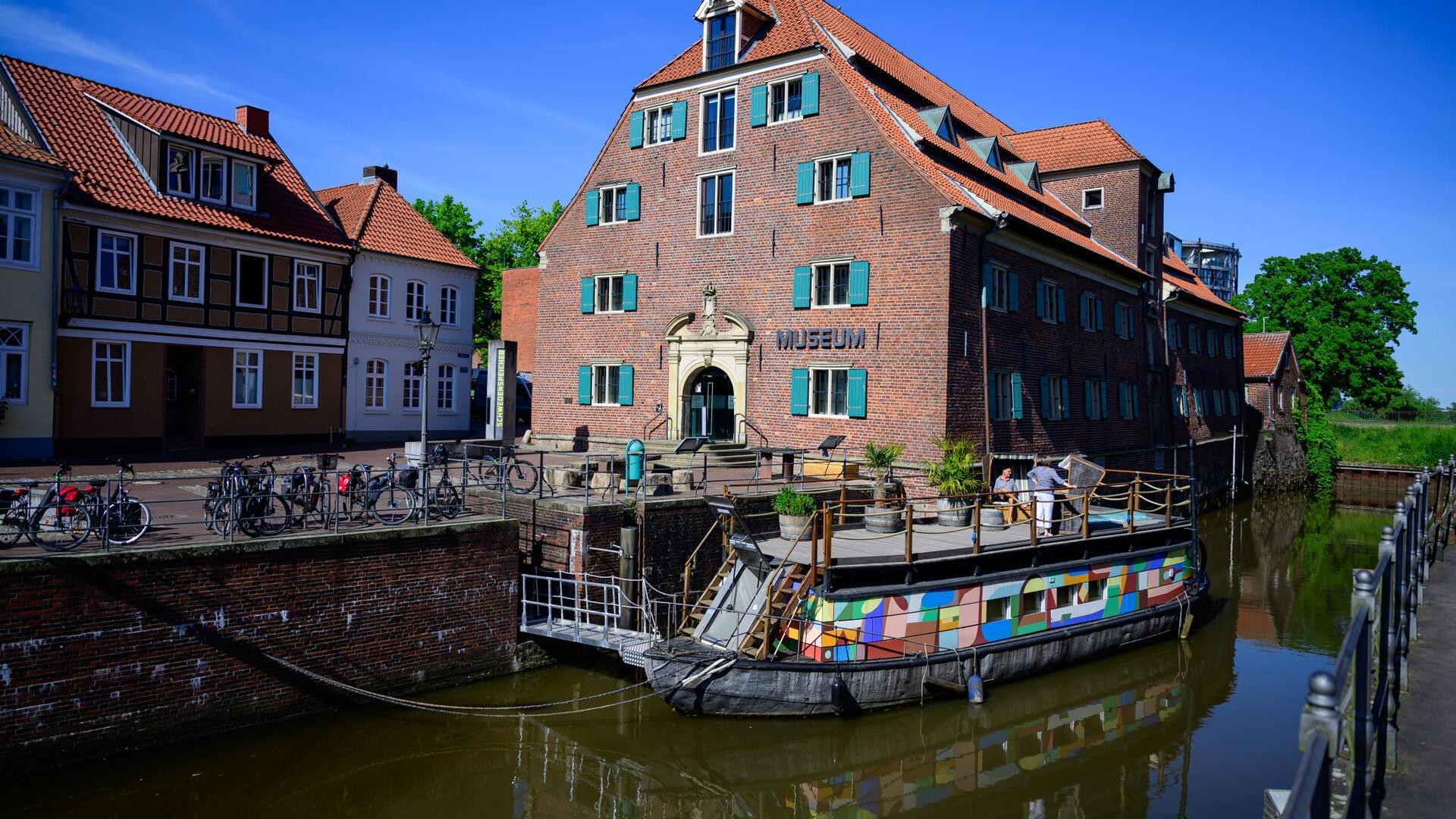 Ein kleines Schiff mit bunten Seitenwänden liegt in einem Hafen. Im Hintergrund sind alte Backsteingebäude zu sehen.  