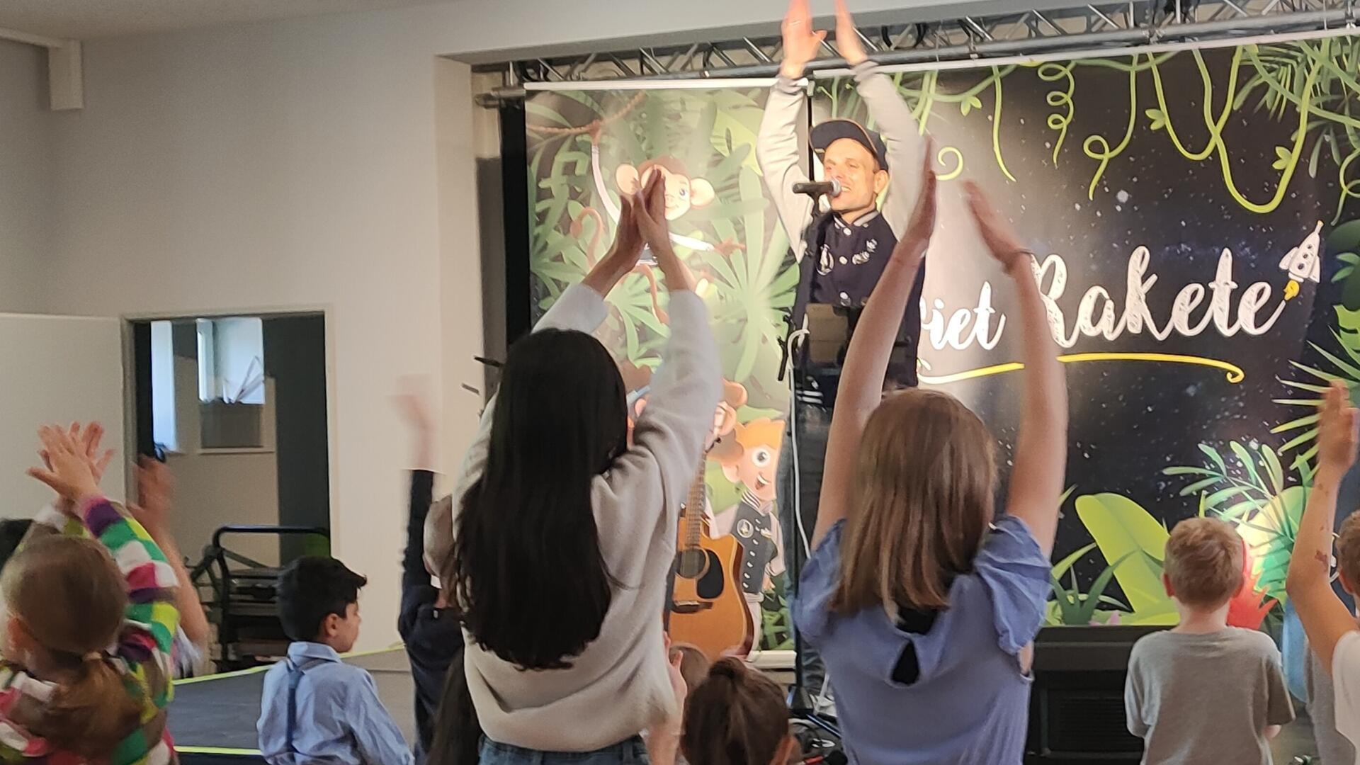 Ein Mann steht auf einer Bühne und hebt die Hände, mehrere Kinder stehen davor.
