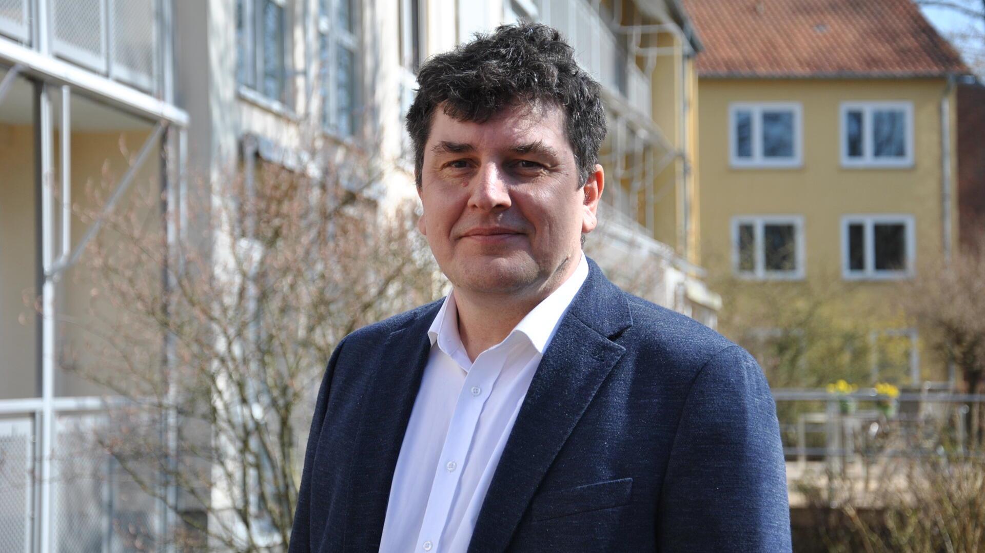 Ein Mann mit kurzen dunklen Locken steht vor einem Haus.