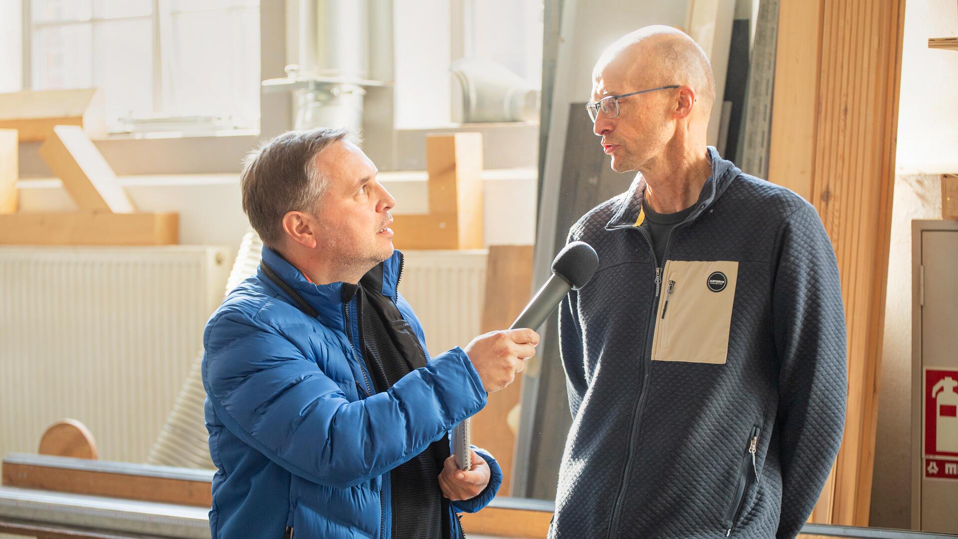ein Reporter mit Mikrofon spricht in einer Holzwerkstatt mit einem Mann mit Brille