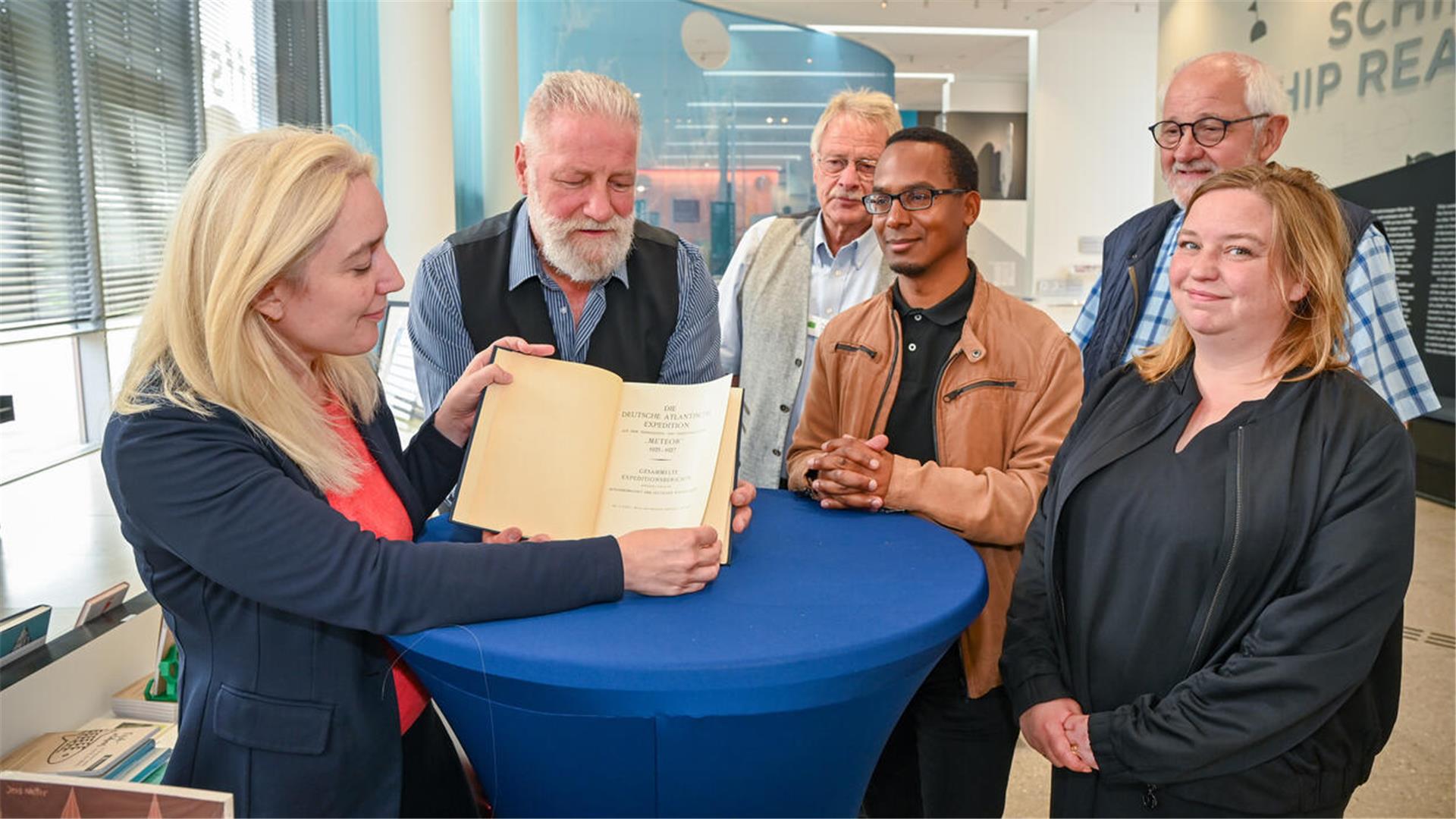 Menschen haben sich um einen Tisch mit blauer Decke versammelt, eine blonde Frau hat ein Buch aufgeschlagen und zeigt es der Kamera. 