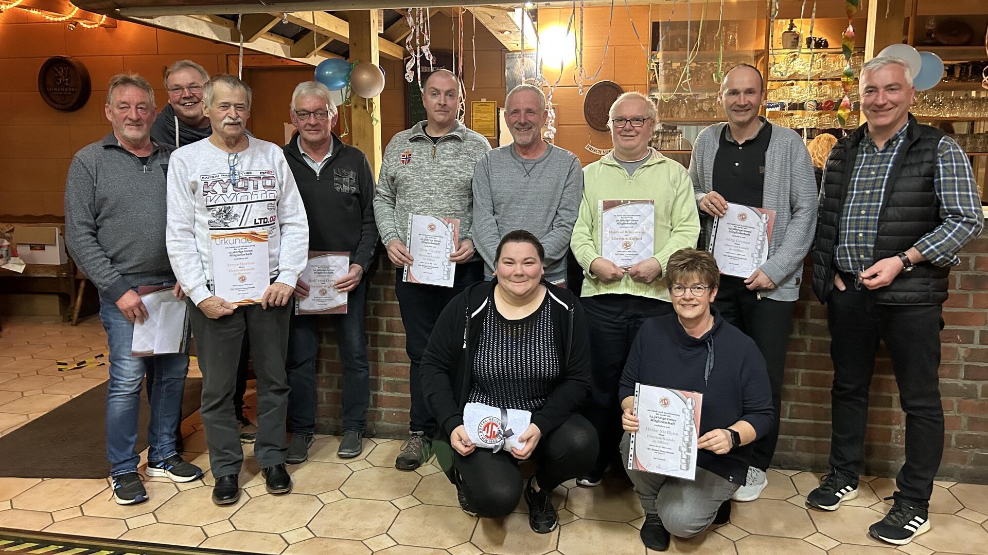 Eine Gruppe von elf Personen, überwiegend Männer, posiert in einem gemütlich dekorierten Raum mit Luftschlangen und Ballons. Mehrere Personen halten Urkunden in den Händen.
