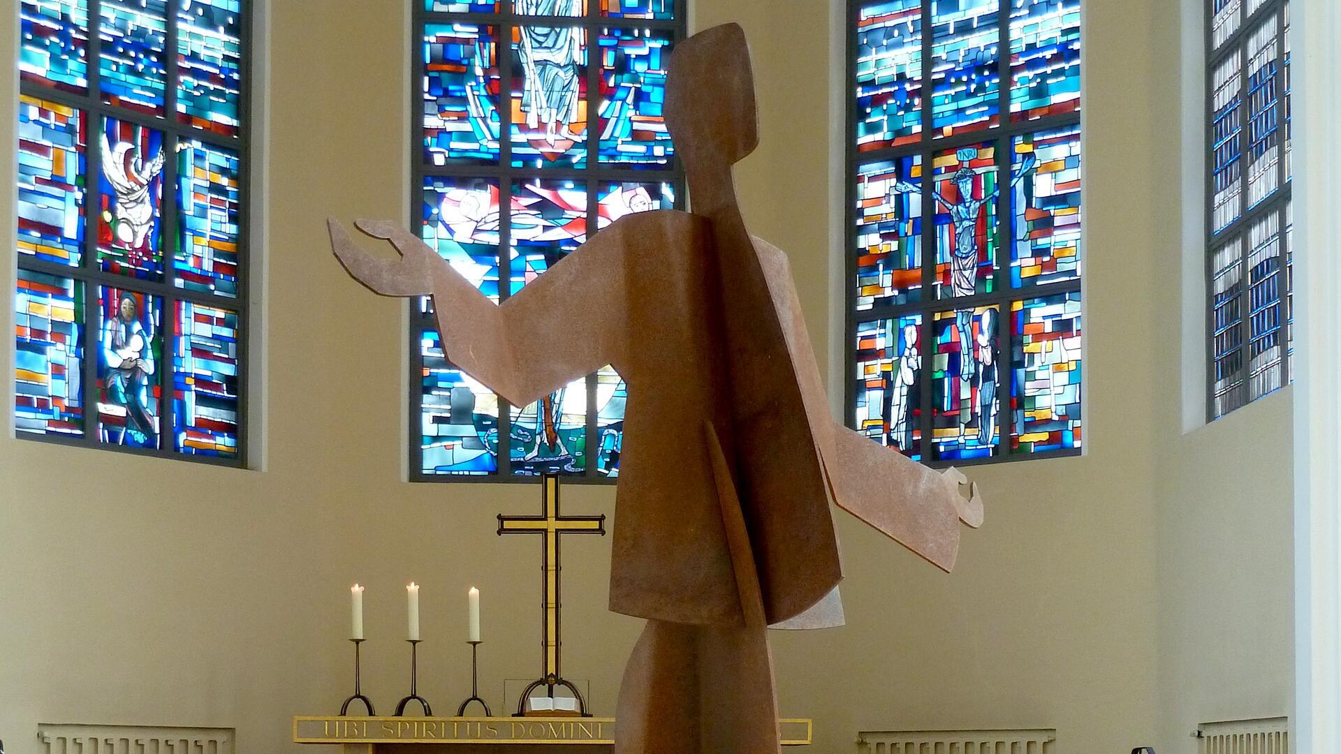 ALT-Text:
Skulptur „Tanzender Christus“ aus Stahl in der Großen Kirche Bremerhaven vor farbigen Kirchenfenstern mit biblischen Motiven und einem Altar mit drei brennenden Kerzen.