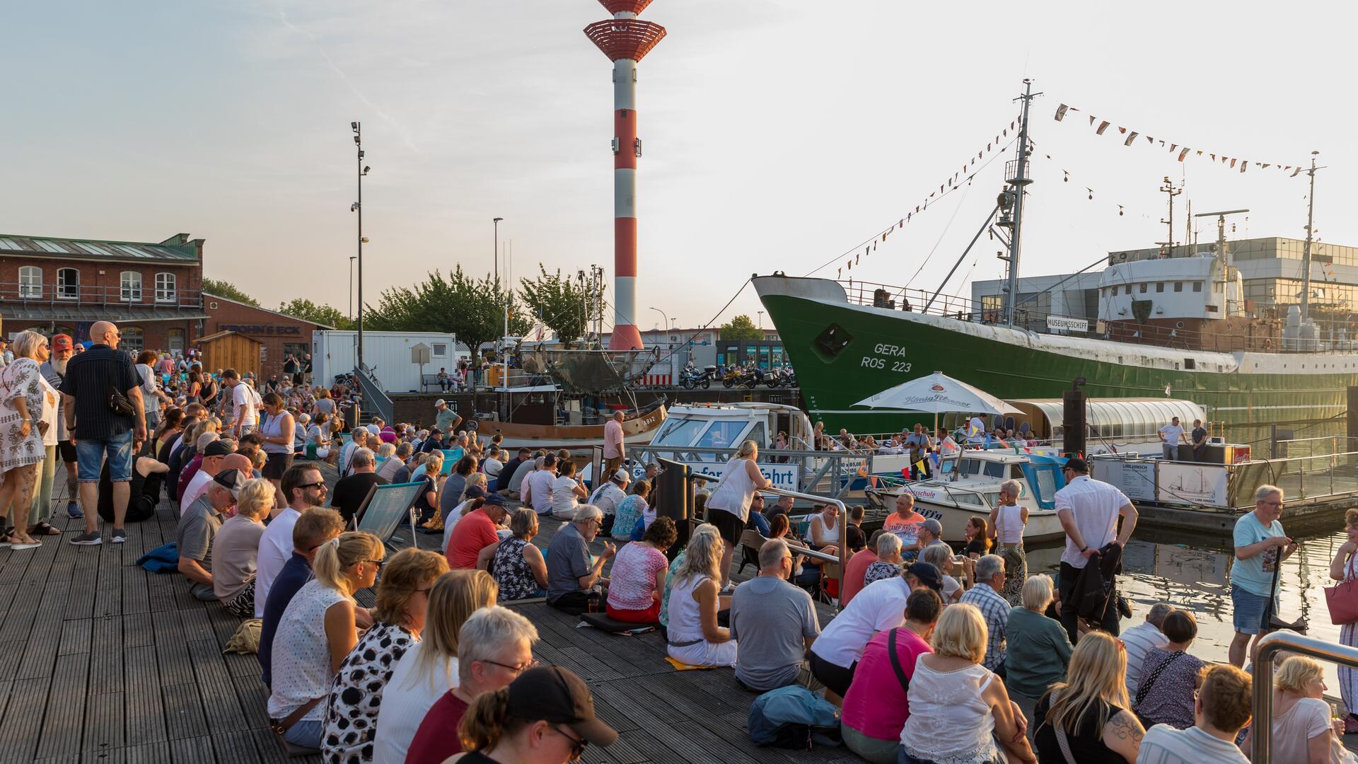 Blick auf den Hafen im Schaufenster Fischereihafen. Zu sehen sind das Museumsschiff "Gera" und das Oberfeuer sowie sitzende Menschen auf der Holzterrasse.