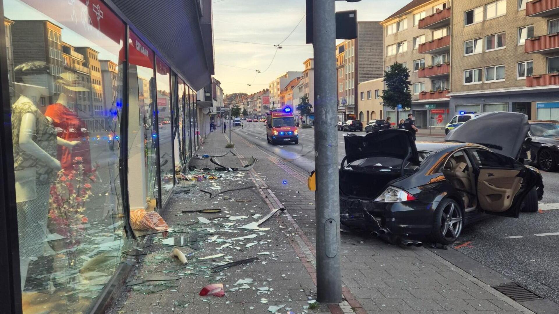 Glasscherben von einem Schaufenster liegen auf einem Fußweg. Daneben steht ein stark beschädigter schwarzer Wagen.