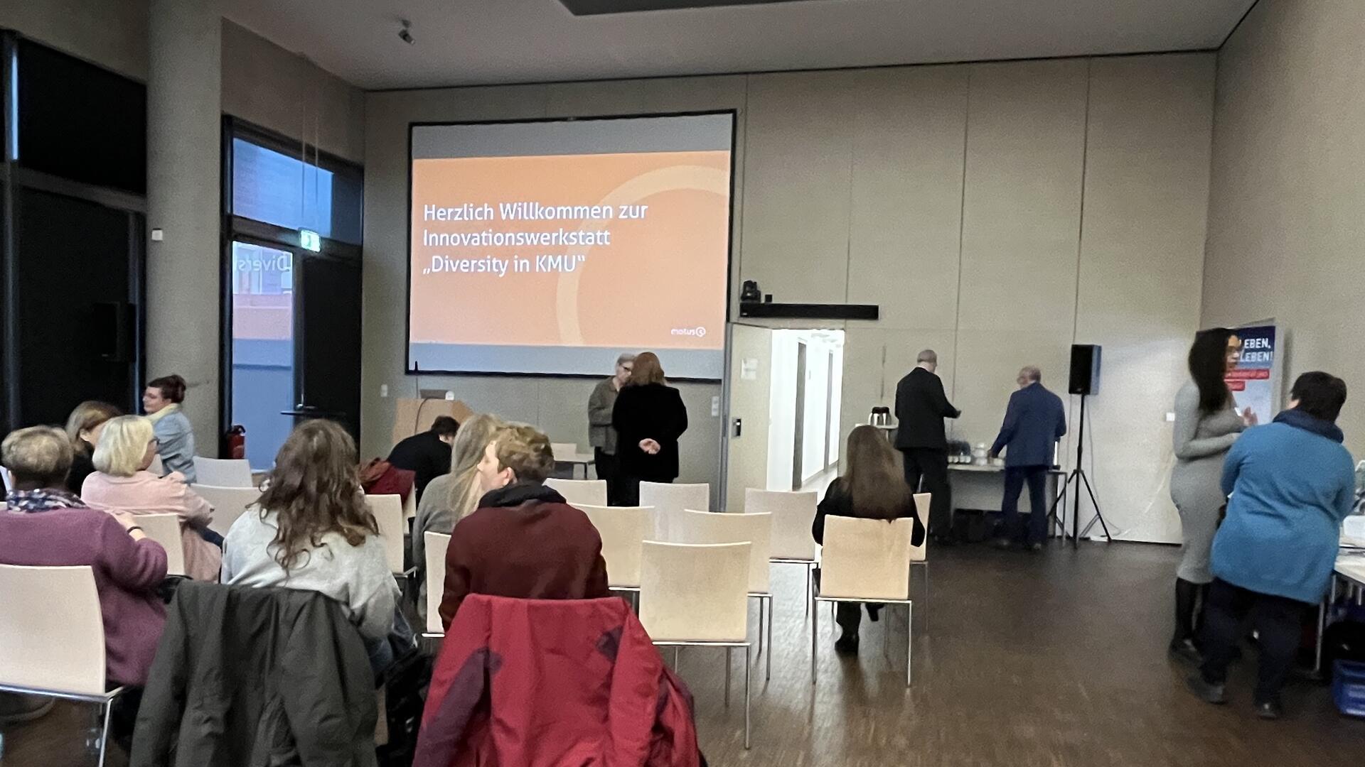 Ein Konferenzraum, in dem mehrere Menschen sitzen und auf einen Bildschirm schauen, auf dem „Herzlich willkommen zur Innovationswerkstatt Diversity in KMU“ steht.