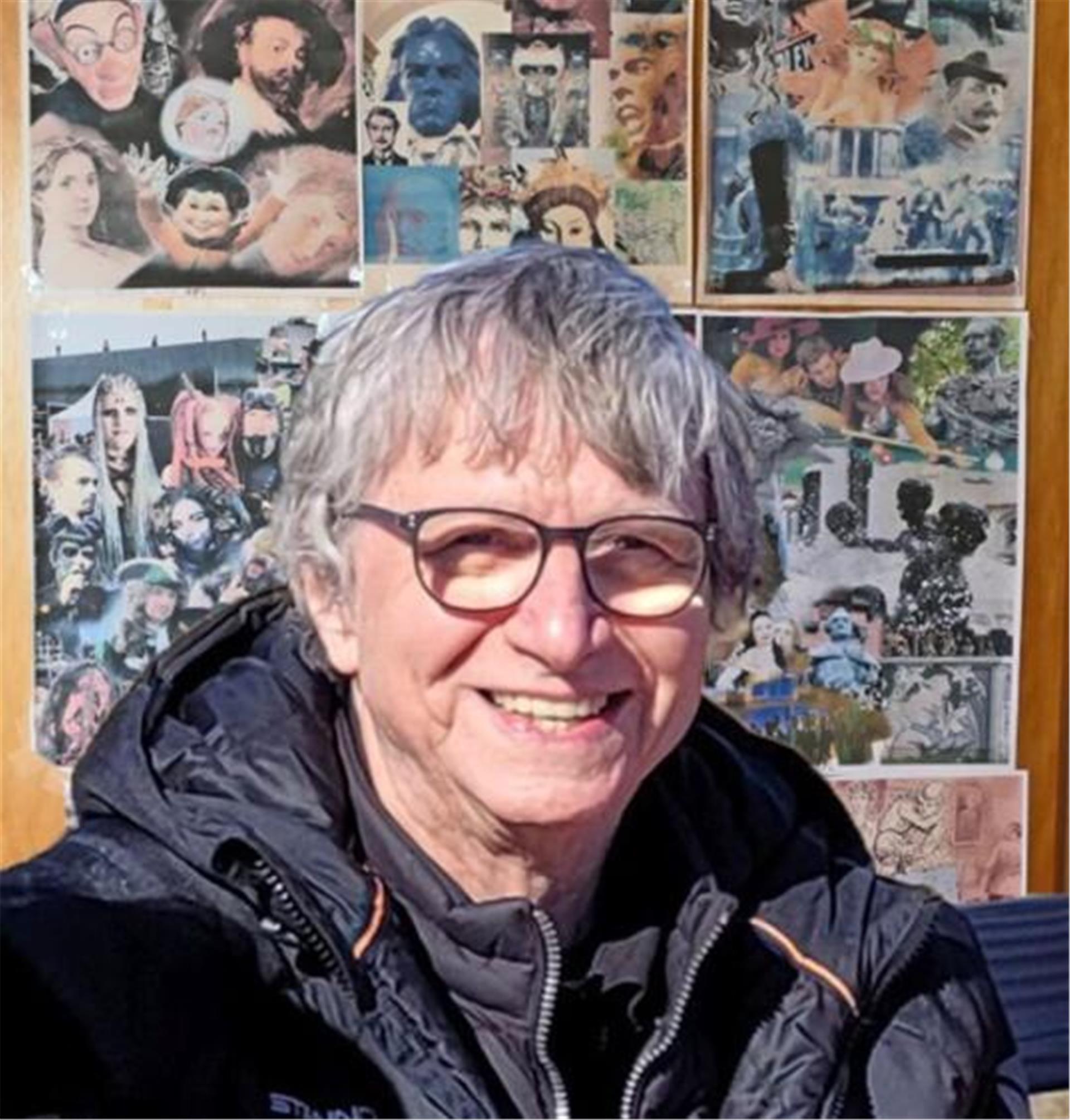 Ein Mann mit grauen Haaren und Brille steht vor einer Wand mit Bildern.