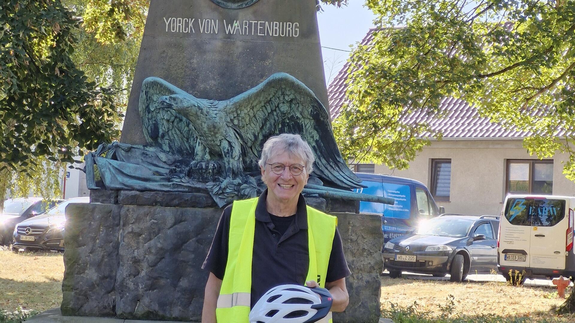 Ein Mann in einer signalgelben Weste und einem Helm in der Hand steht vor einem Denkmal.