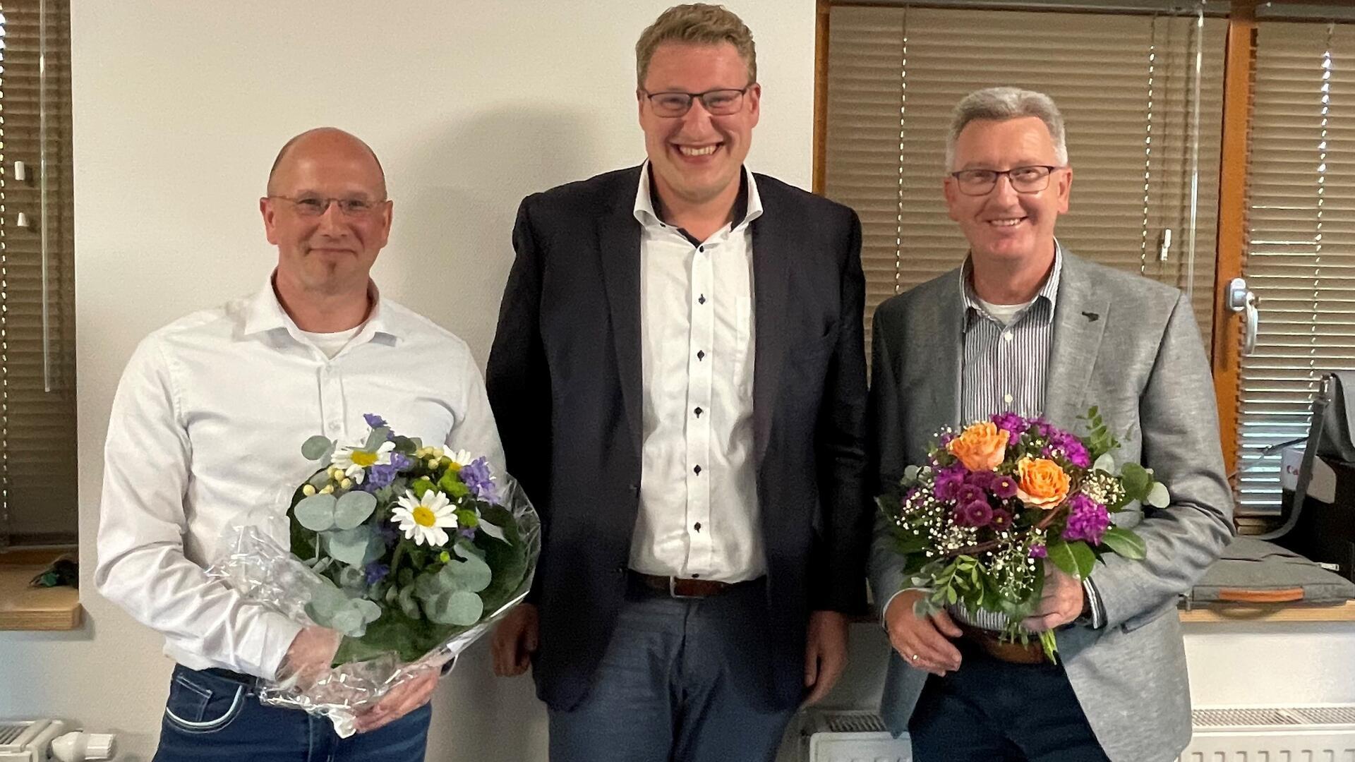 Drei Männer, zwei von ihnen halten je einen Blumenstrauß in den Händen.