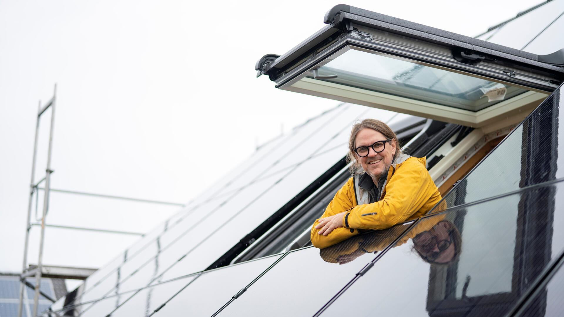 Ein Mann mit Brille und gelber Jacke lehnt sich lächelnd aus einem geöffneten Dachfenster. Das Dach ist mit glänzenden Solarpaneelen bedeckt. Im Hintergrund ist ein Baugerüst zu sehen.