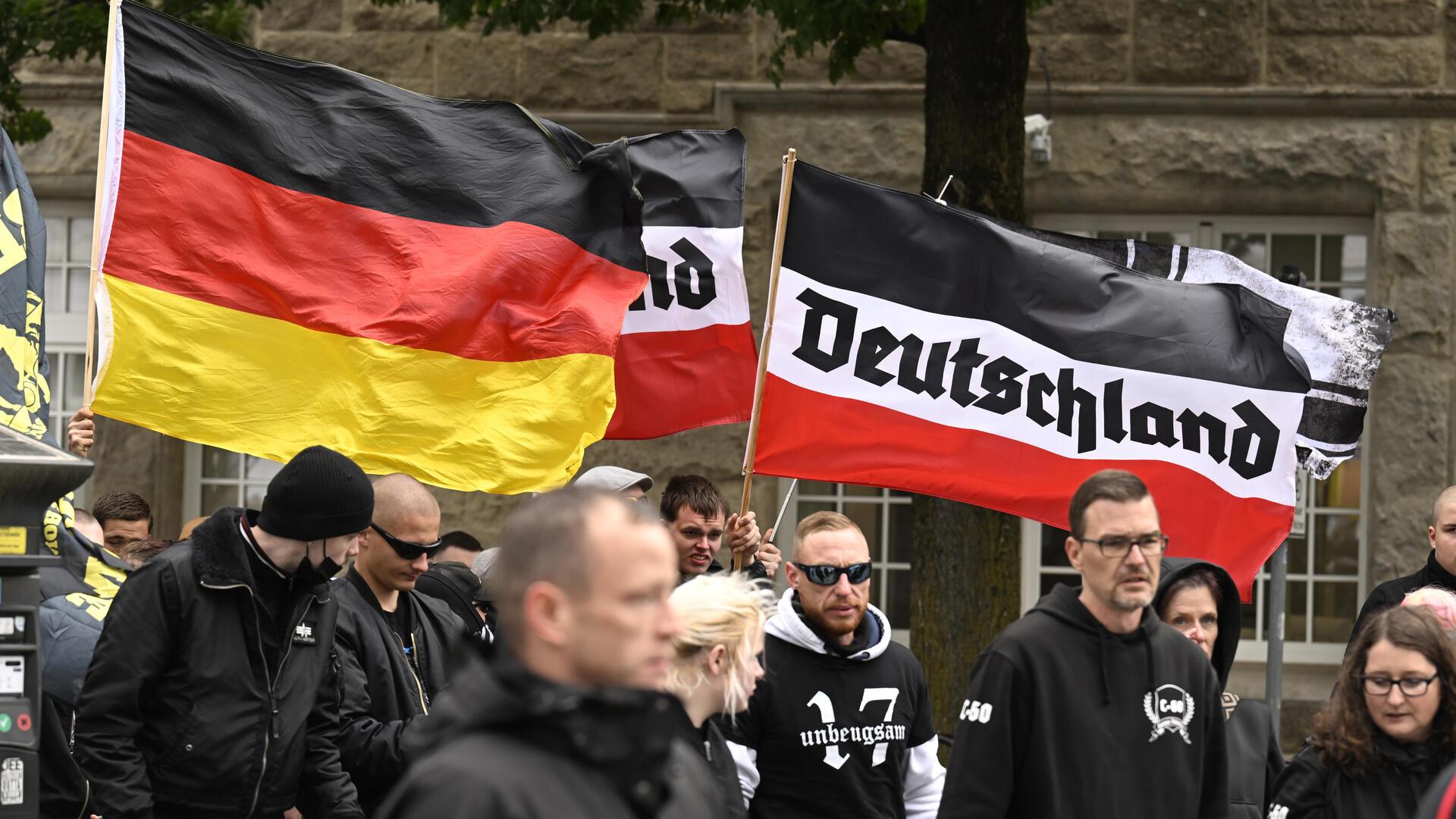 Teilnehmer einer rechtsextremen Demonstration mit schwarz-weiß-roter Flagge mit der Aufschrift „Deutschland“ und einer Deutschlandflagge; mehrere Personen in dunkler Kleidung, teils mit Sonnenbrillen oder Kapuzen, stehen eng beieinander.